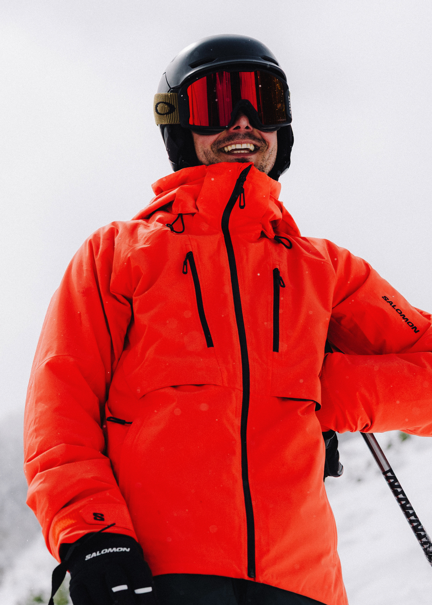 Man in bright orange ski jacket and helmet, holding skis, smiles against a snowy backdrop.