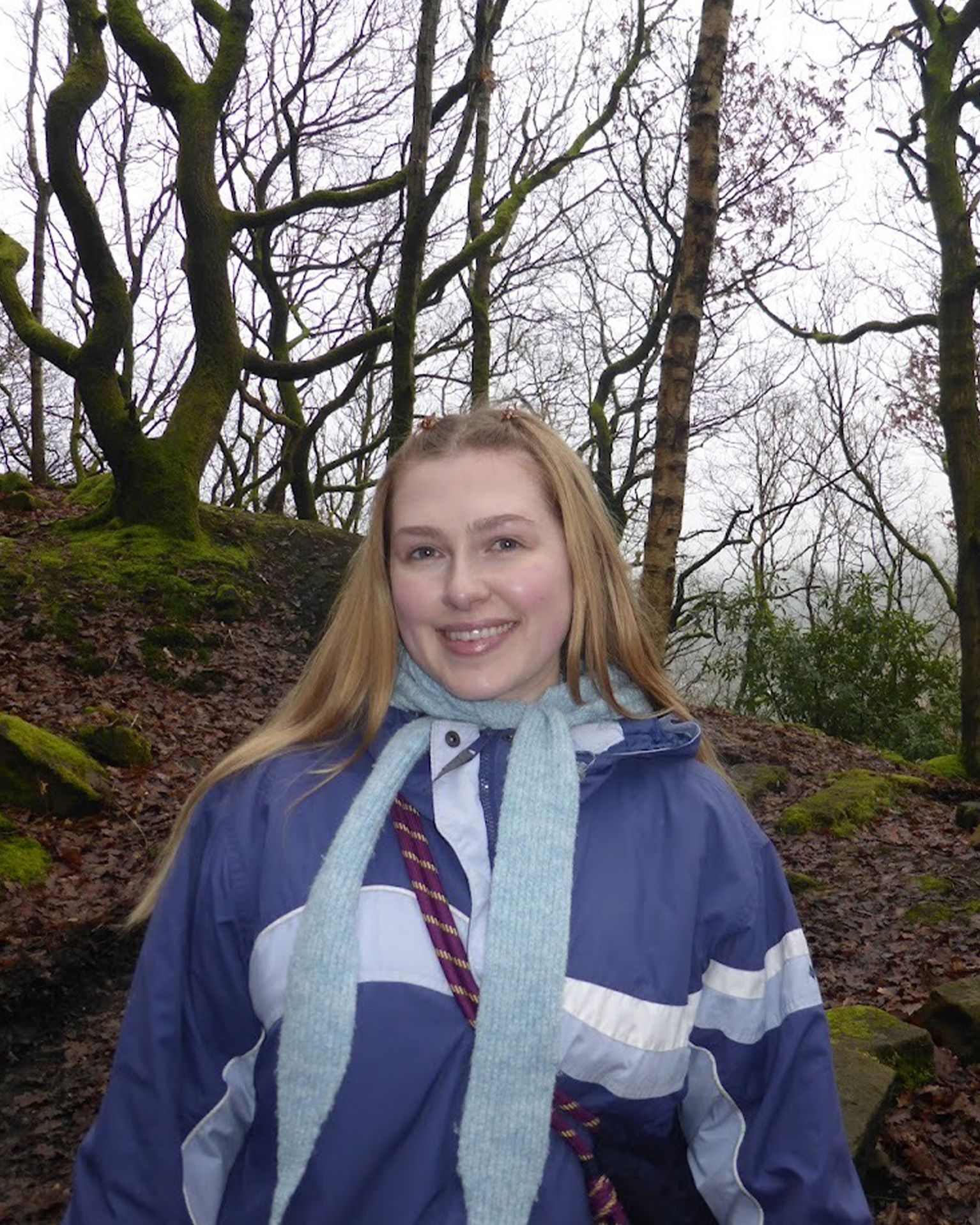 Person smiling while on a woodland trail, wearing a blue jacket and scarf. Leaf-strewn path and moss-covered trees in the background.