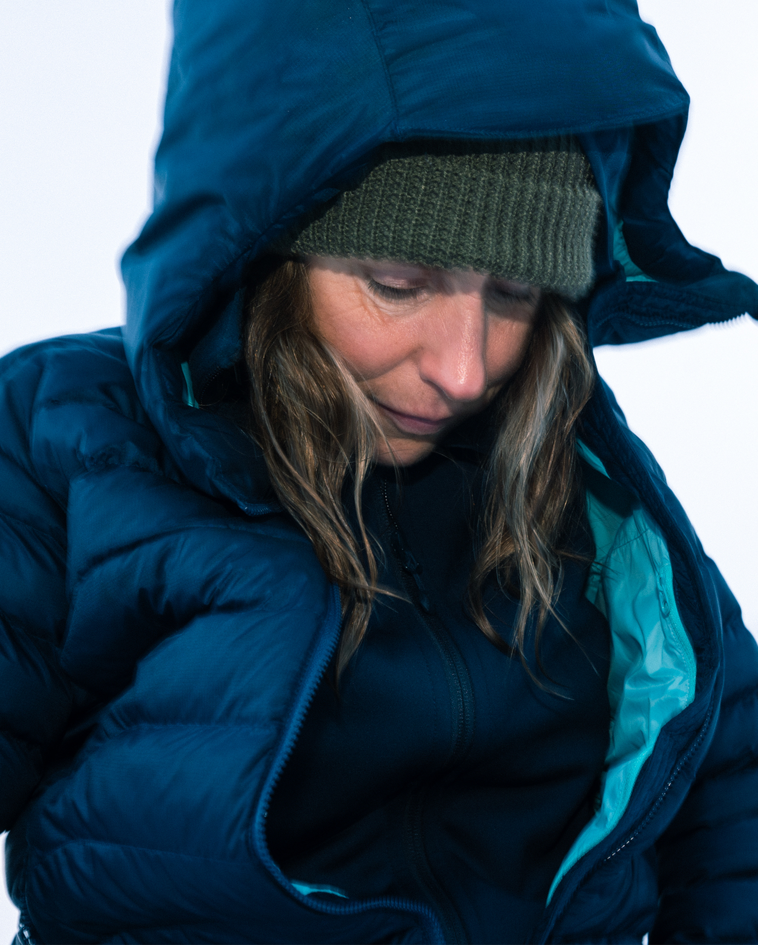 A person wearing a blue hooded jacket and green knit hat looks down, pictured against a plain background.