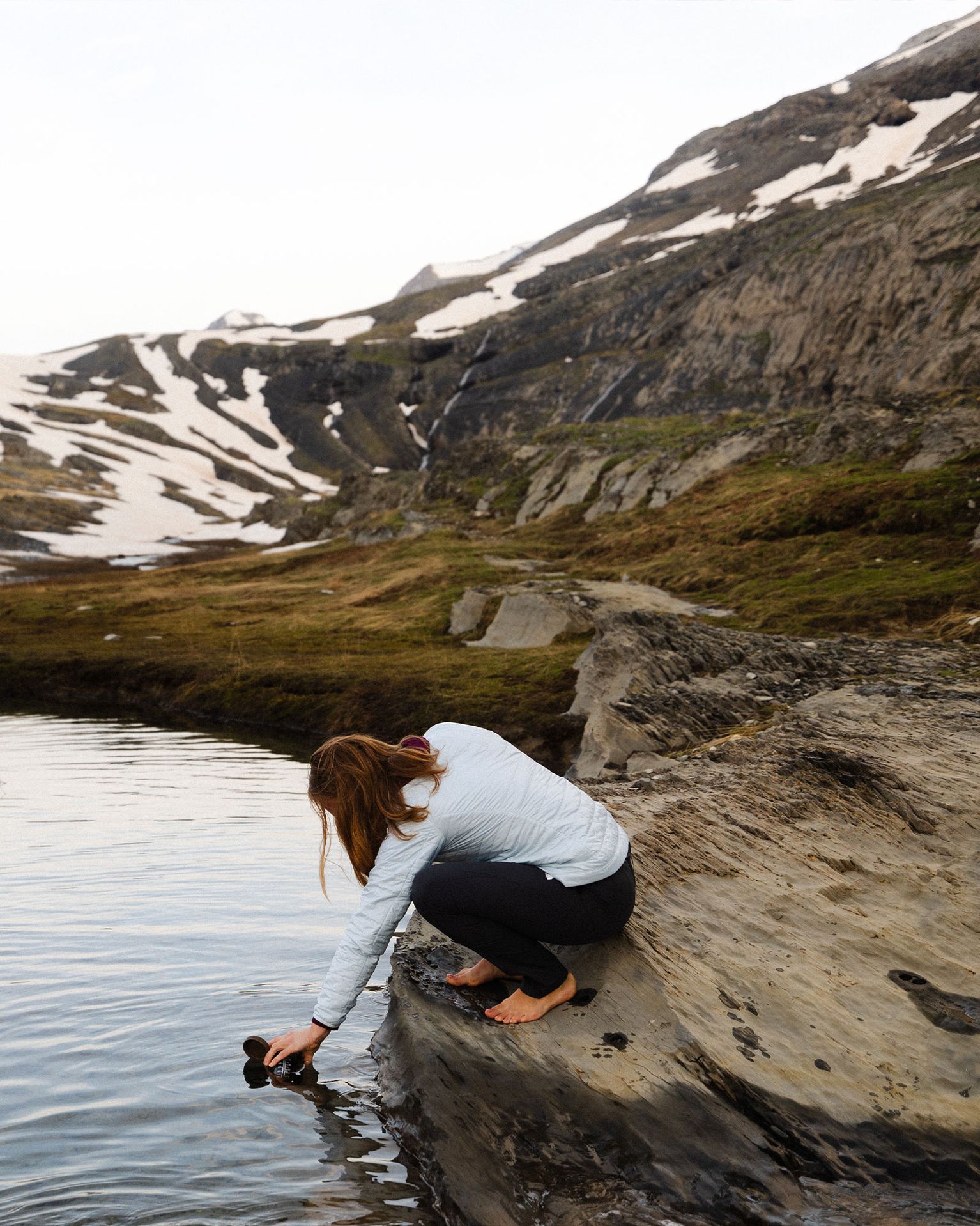 Person kneels on rocky shore, filling a cup from a mountain stream, surrounded by rugged, snow-dappled peaks.