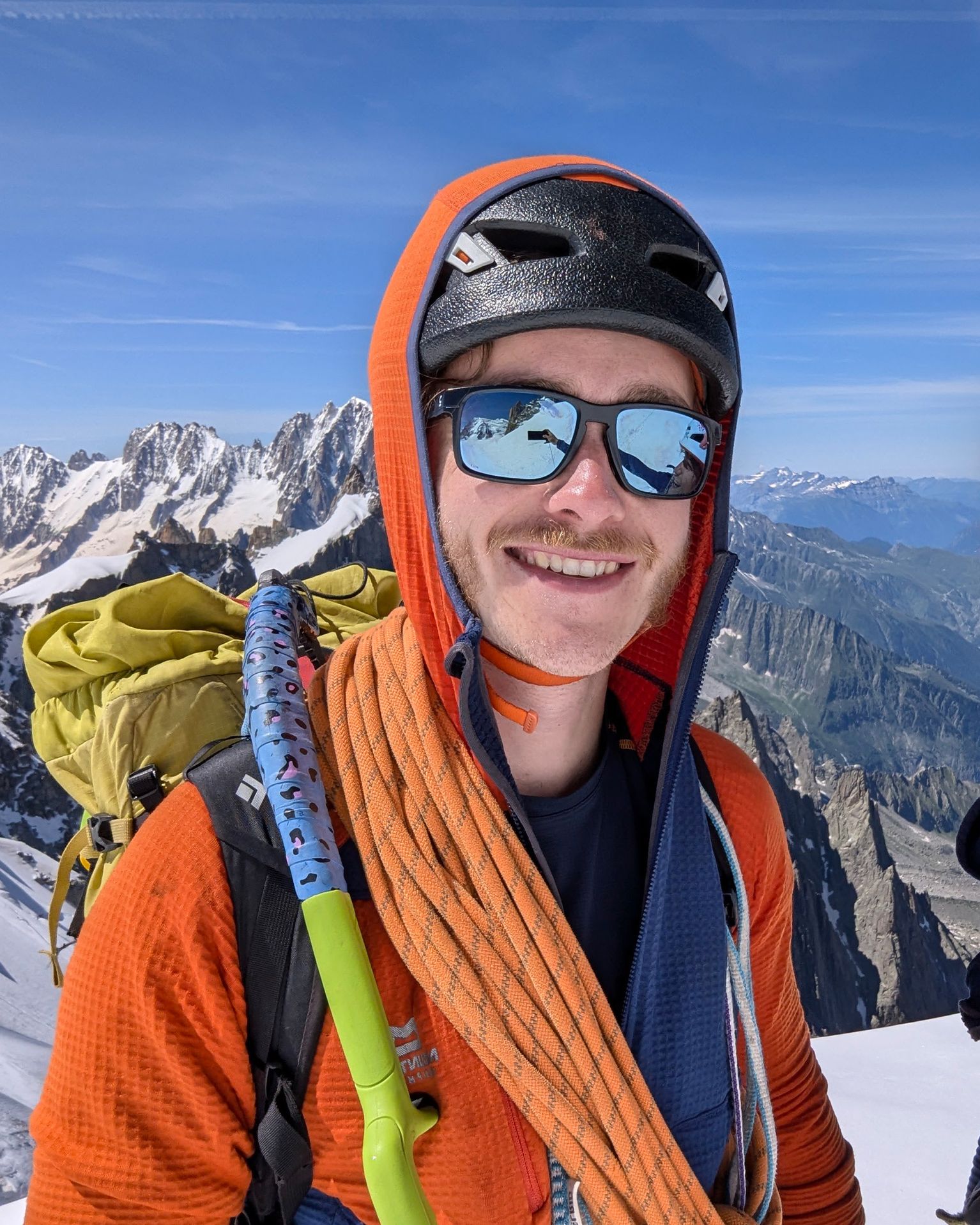 Man on snowy mountain in orange jacket and helmet, wearing sunglasses, with climbing gear and rope, snowcapped peaks in the background.