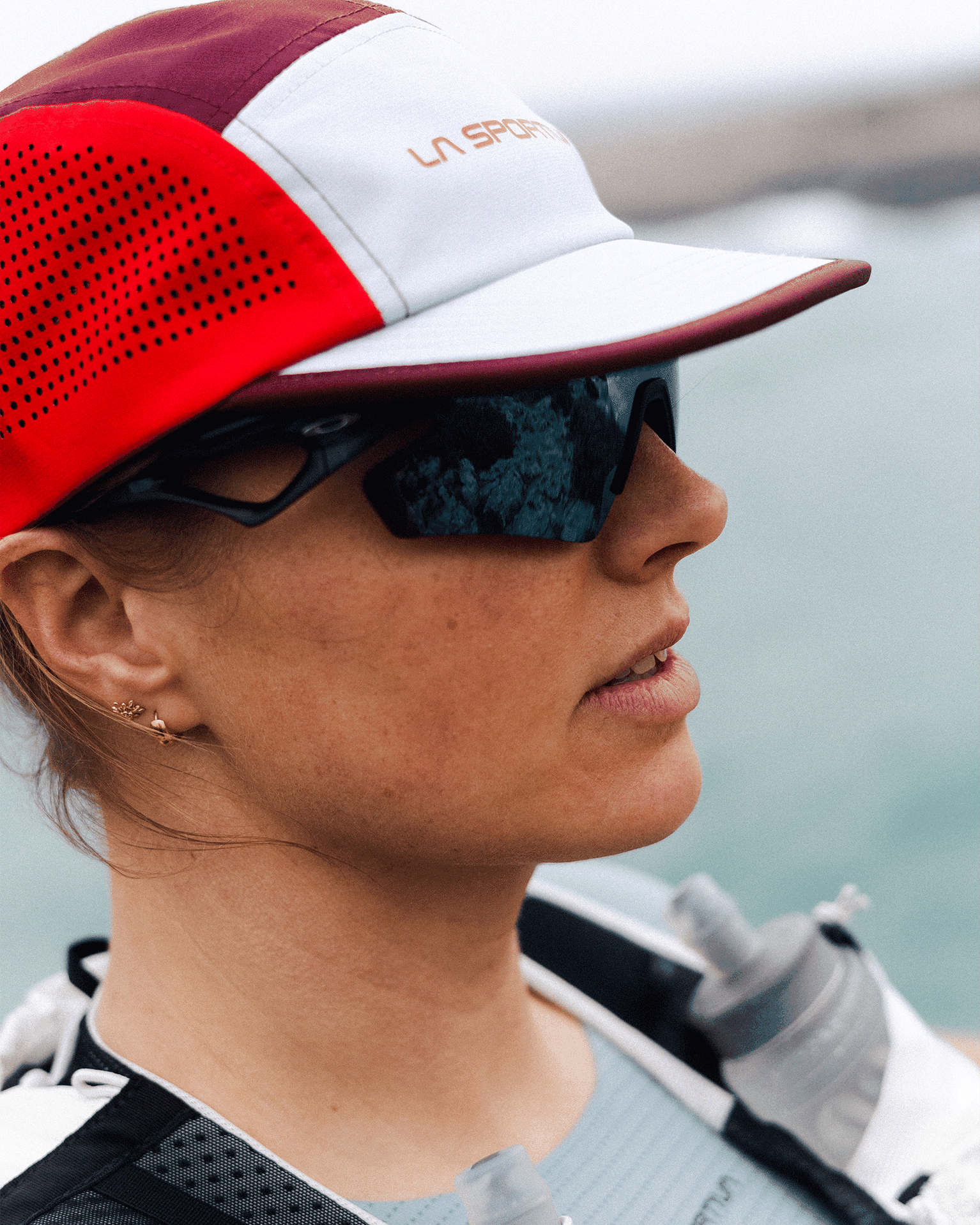 Woman in a red and white cap wearing mirrored sunglasses and running gear, standing near a body of water with a focused expression.