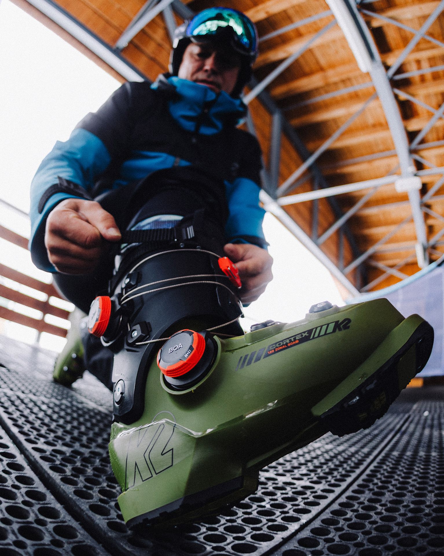 Person adjusting green ski boots with red dials, wearing a blue and black jacket, under a wooden ceiling structure.