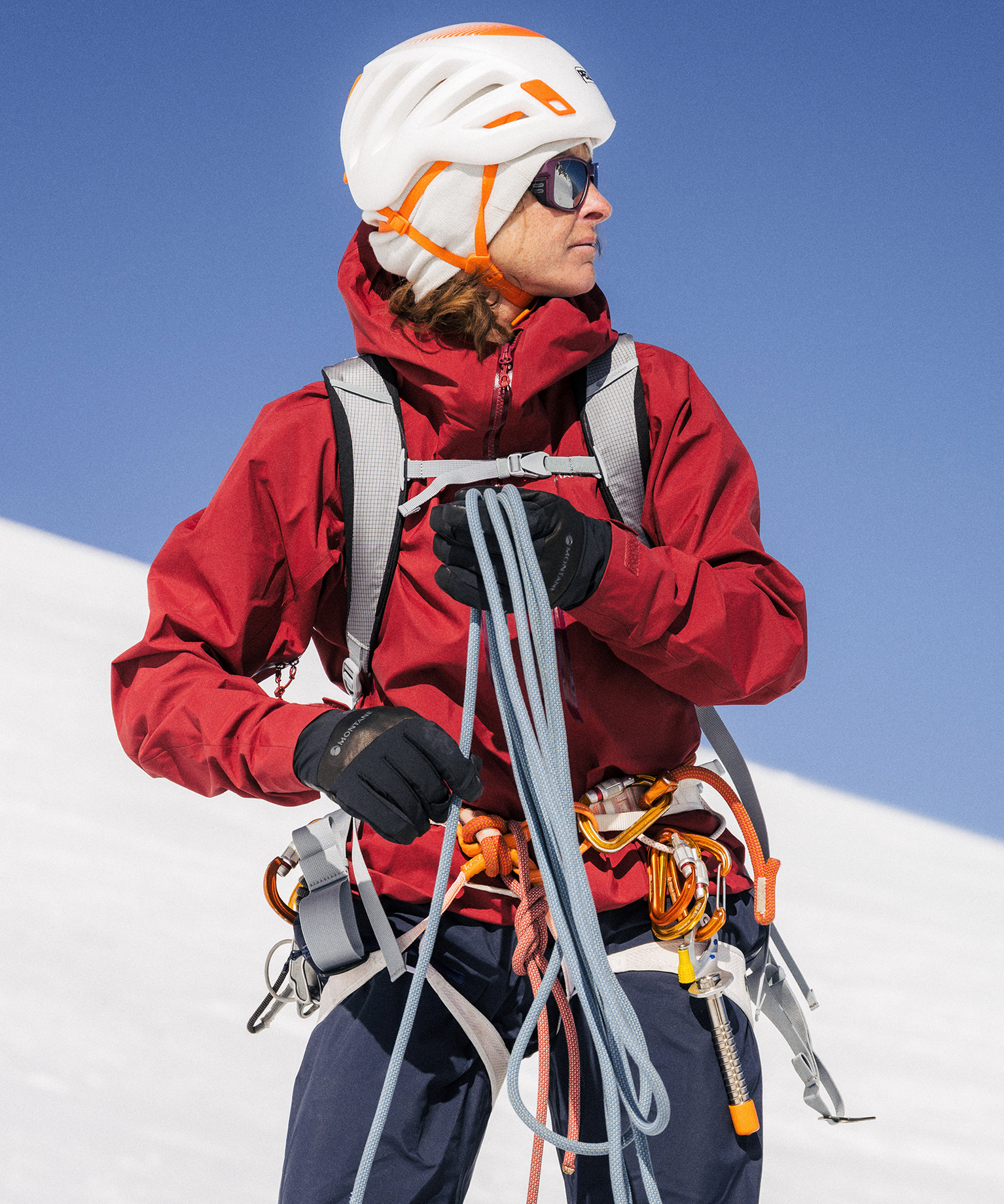 A climber in a red jacket and white helmet holds climbing ropes on a snowy mountain slope under a clear blue sky.