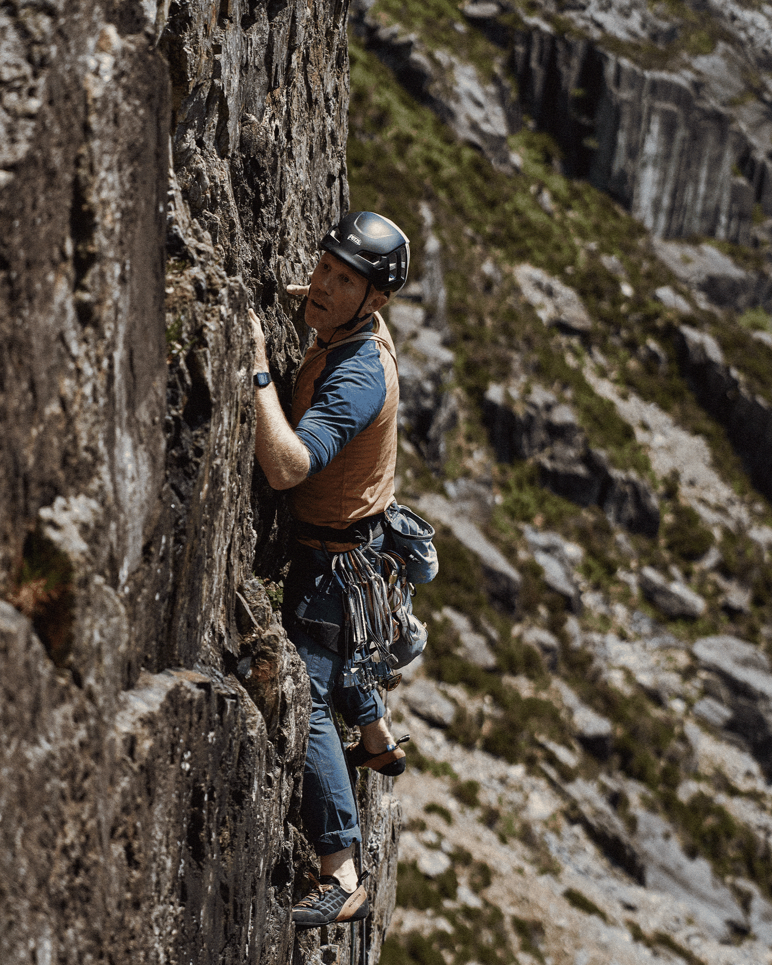 Rock climber in a yellow jacket and helmet scaling a rugged cliff face, equipped with gear, against a mountainous backdrop.