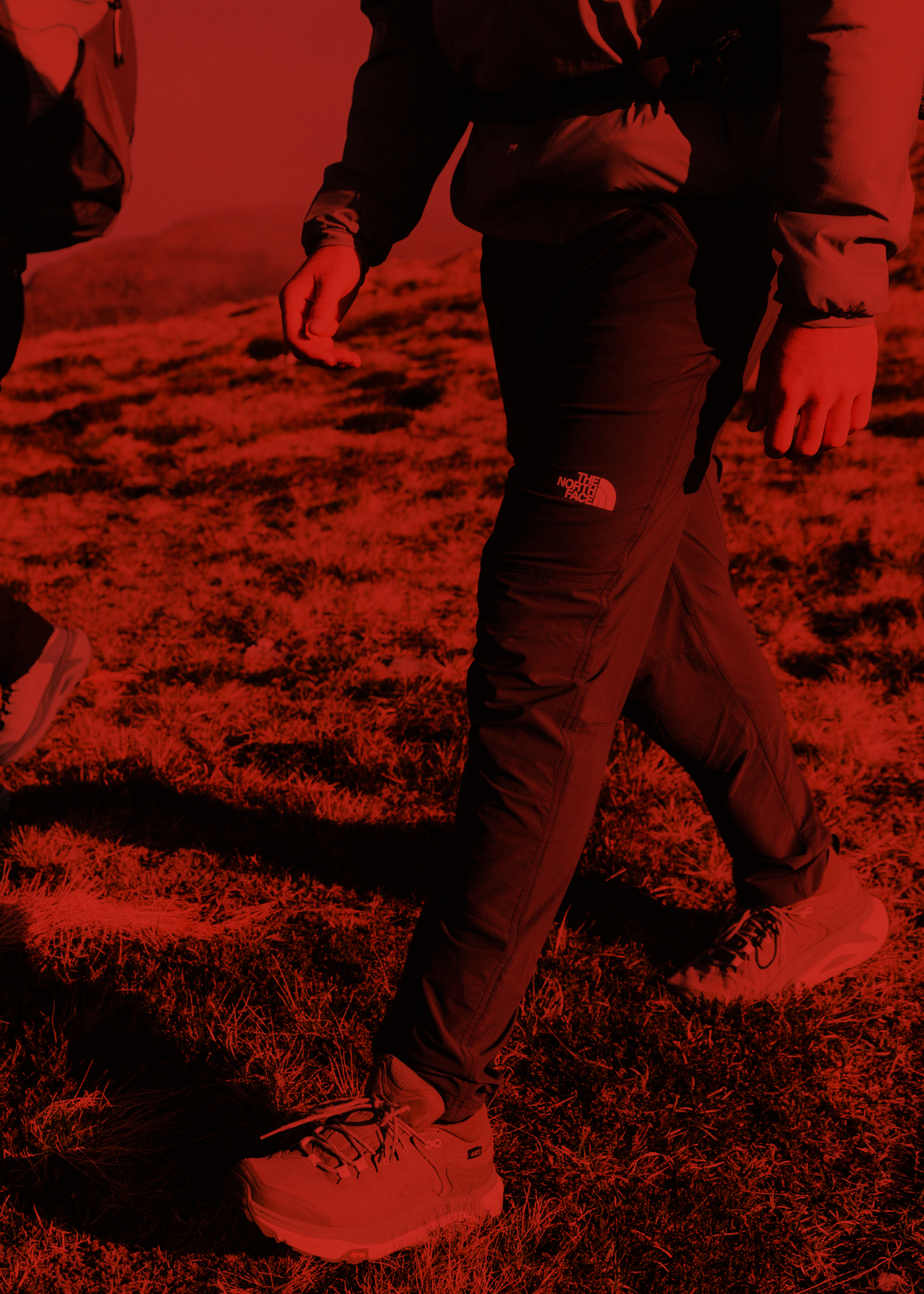 Two hikers wearing outdoor clothing, walking on grassy terrain, with the image in a red tint.