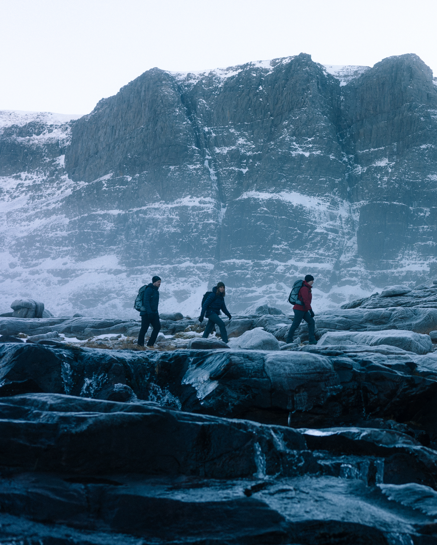 Three hikers traverse icy rocky terrain beneath towering snow-dusted mountains. Cold, crisp atmosphere under a wide, pale sky.