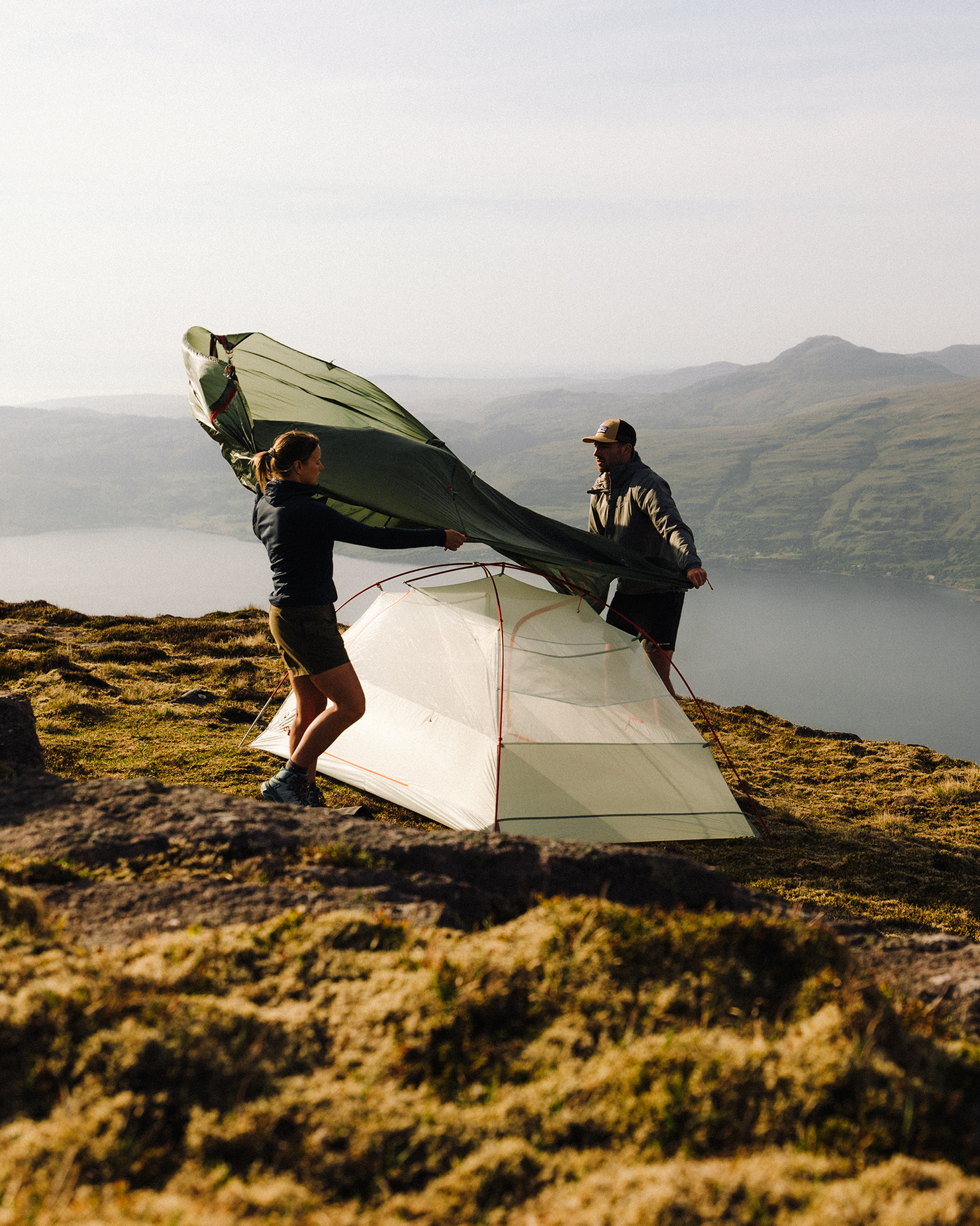 Two people assembling a tent on a mountain, overlooking a scenic view with hills and a body of water in the background.