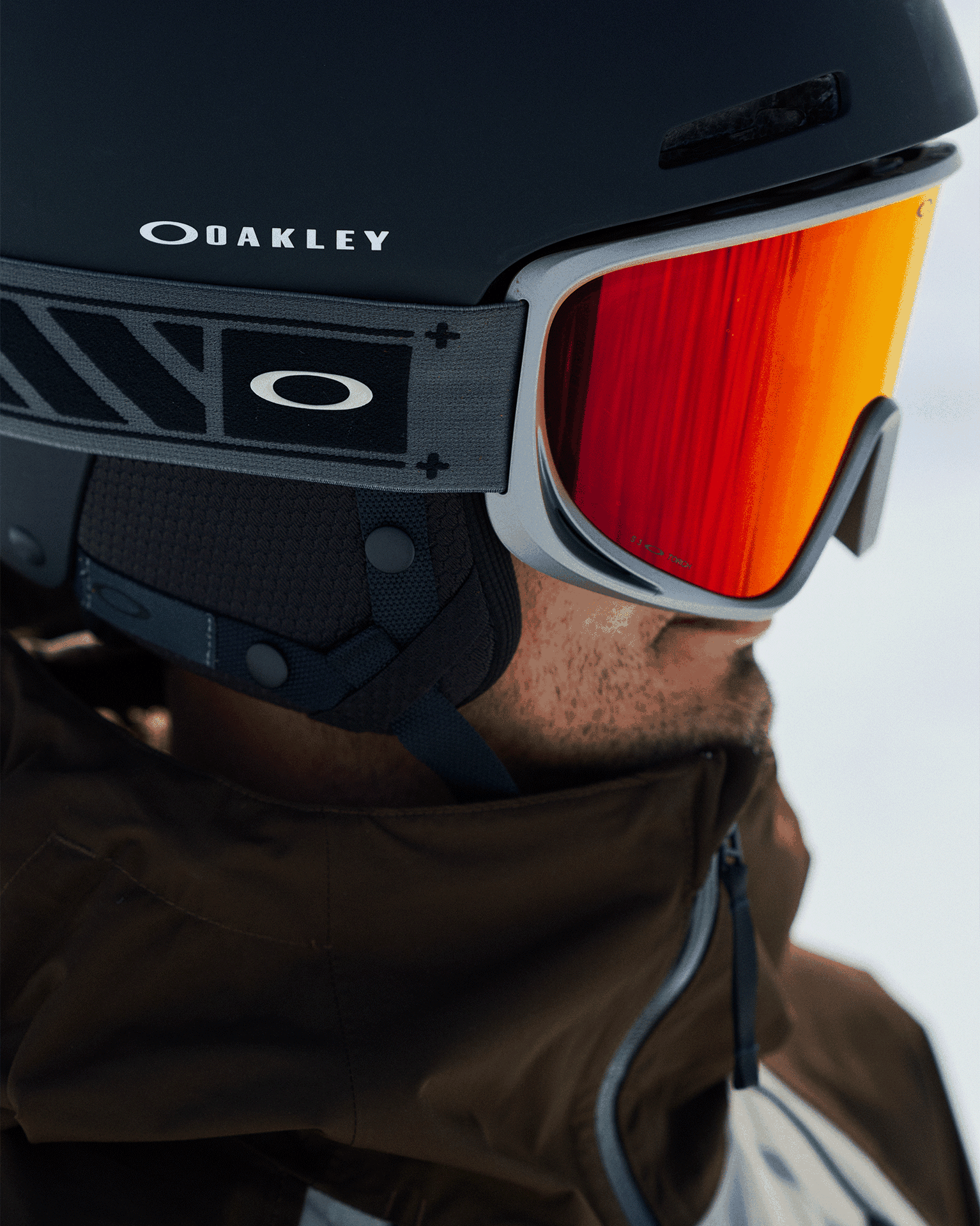 Person wearing black ski helmet and red-tinted goggles looking into distance, brown jacket visible, close up on helmet and goggles detail.