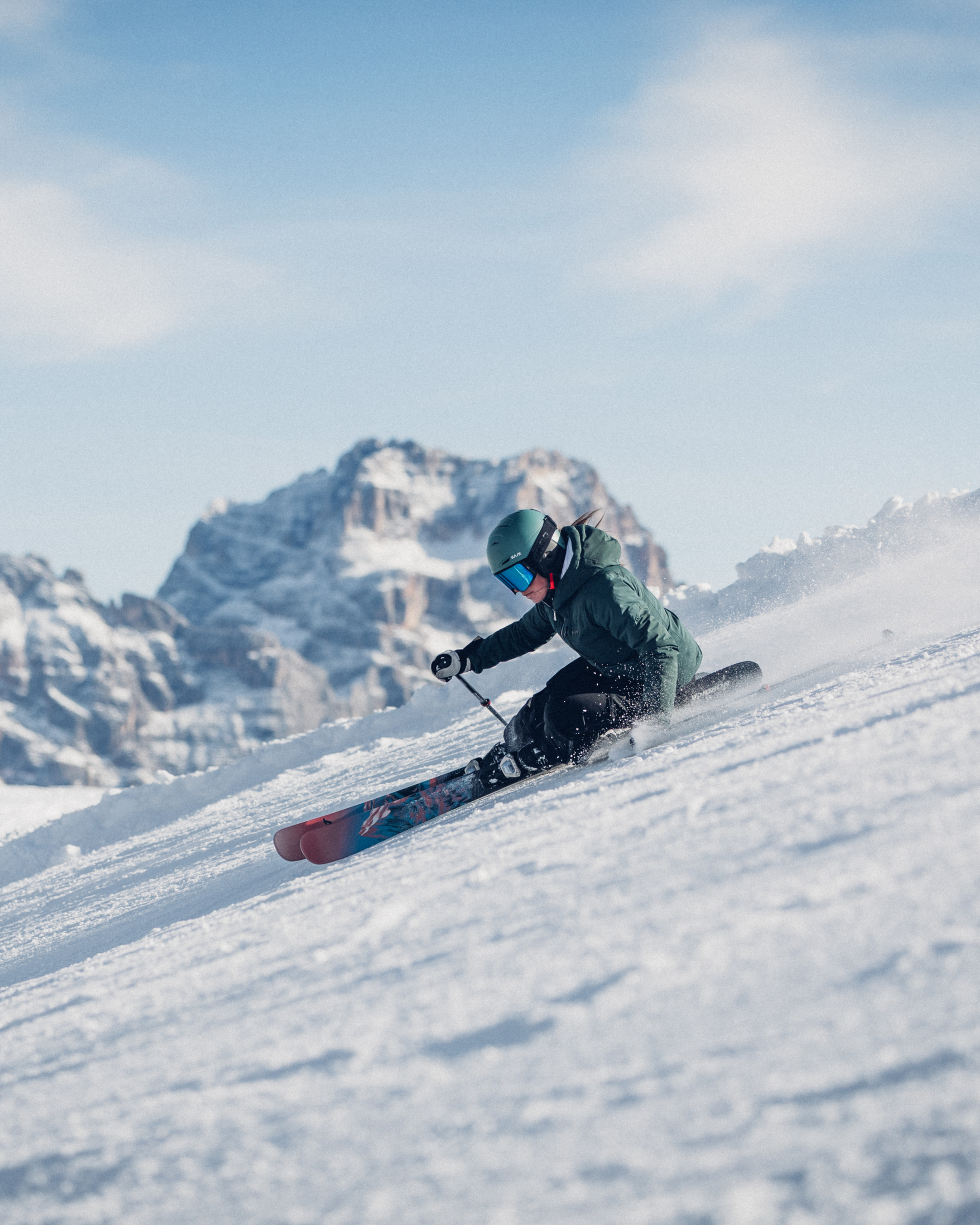 Skier in green jacket and helmet carving down a snowy slope with a mountain peak in the background.