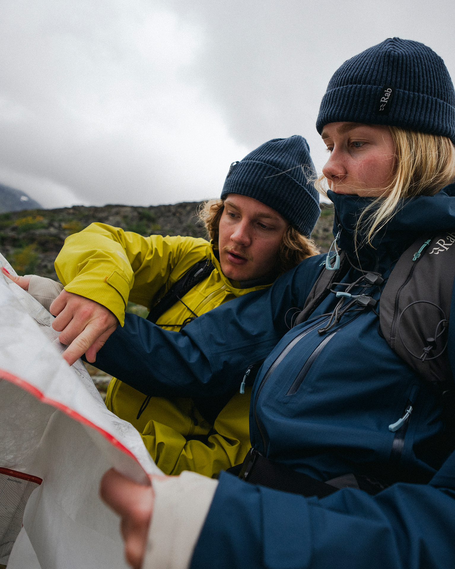 Two people in outdoor gear study a map against a cloudy mountain backdrop. They appear focused and prepared for hiking.