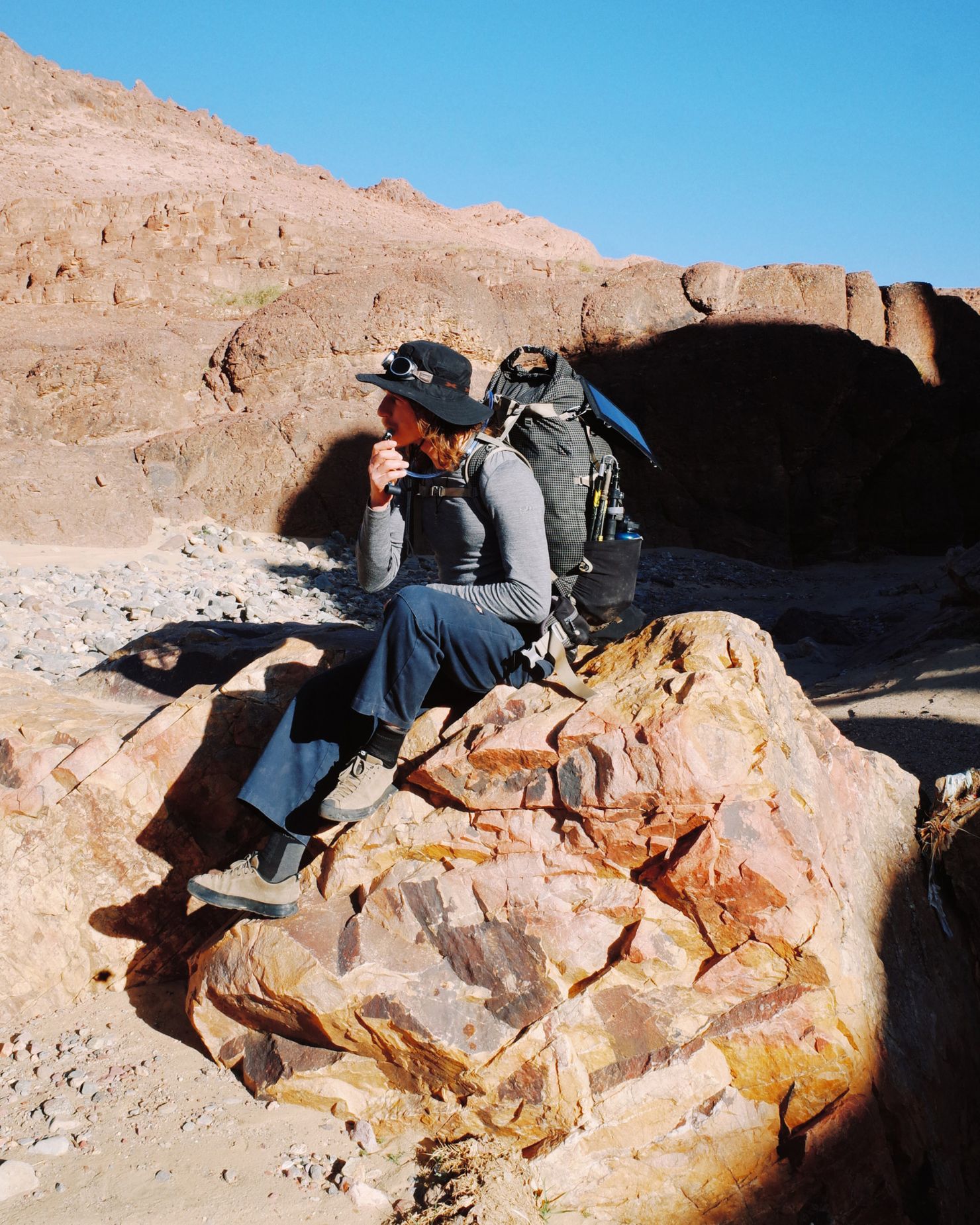 Person wearing hiking gear, seated on a large rock in a rugged desert landscape, with a backpack, under clear blue skies.