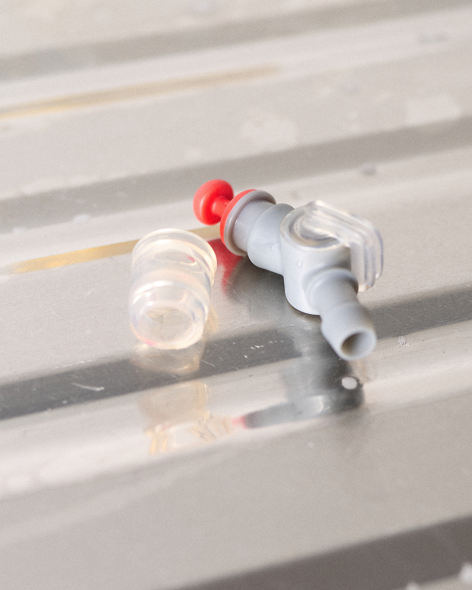 Close-up of a gray plastic valve with a transparent cap and red stopper, lying on a reflective metallic surface.
