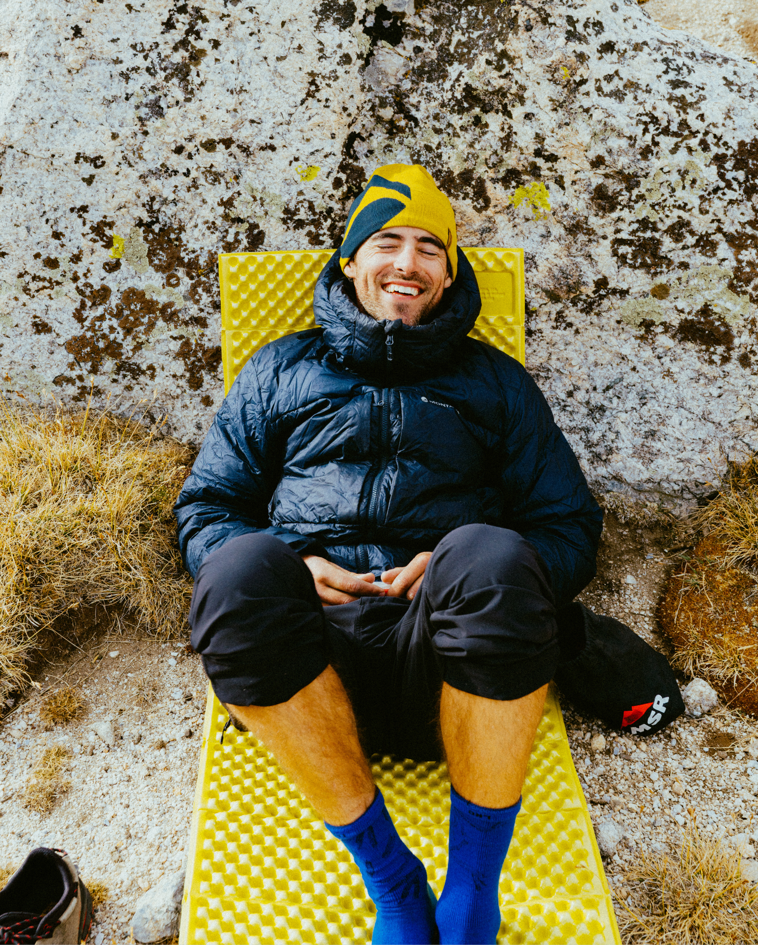 Man in a black jacket and yellow beanie relaxing on a yellow mat against a rocky terrain, smiling.