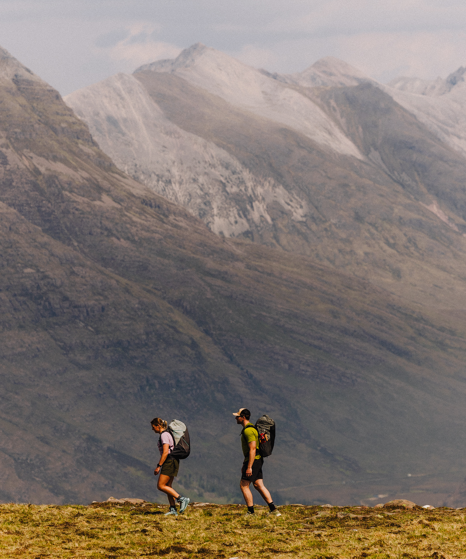 Two hikers with backpacks walk across a grassy terrain with towering, rocky mountains in the background under a cloudy sky.