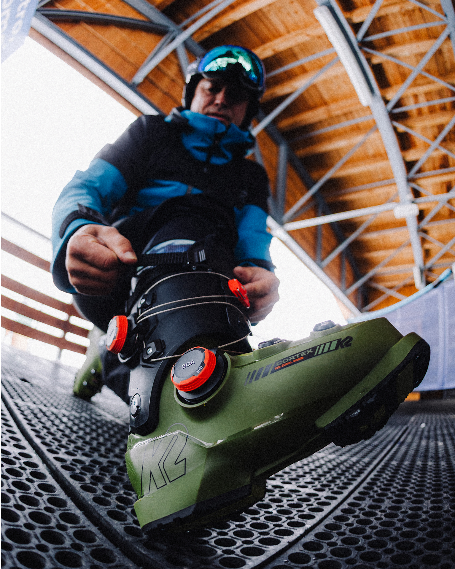 Person fastening green ski boots on a textured floor, wearing a blue and black jacket, with a wooden ceiling and metal beams above.