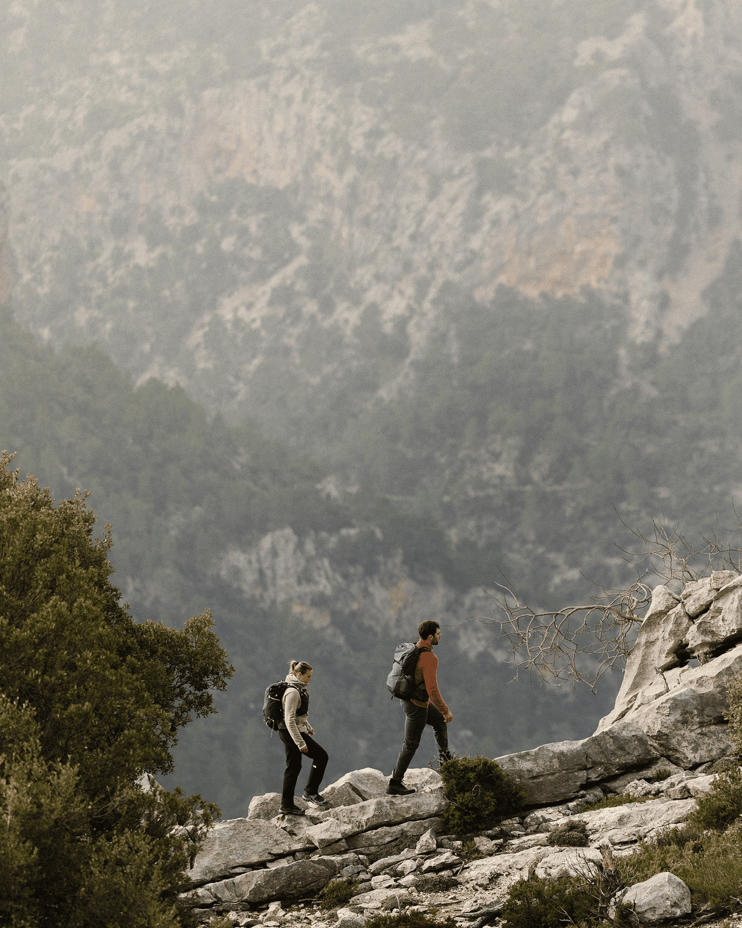 Two people hiking up a rocky mountain trail, surrounded by a misty backdrop of cliffs and trees.