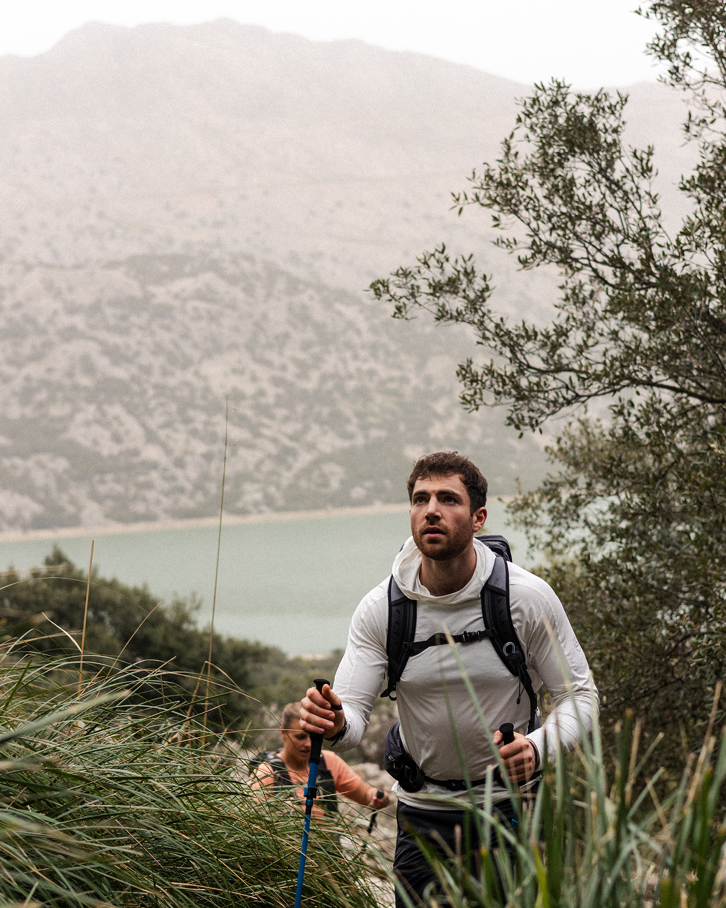 Man hiking through lush greenery with a backpack and trekking poles, misty mountains in the background, conveying adventure and endurance.