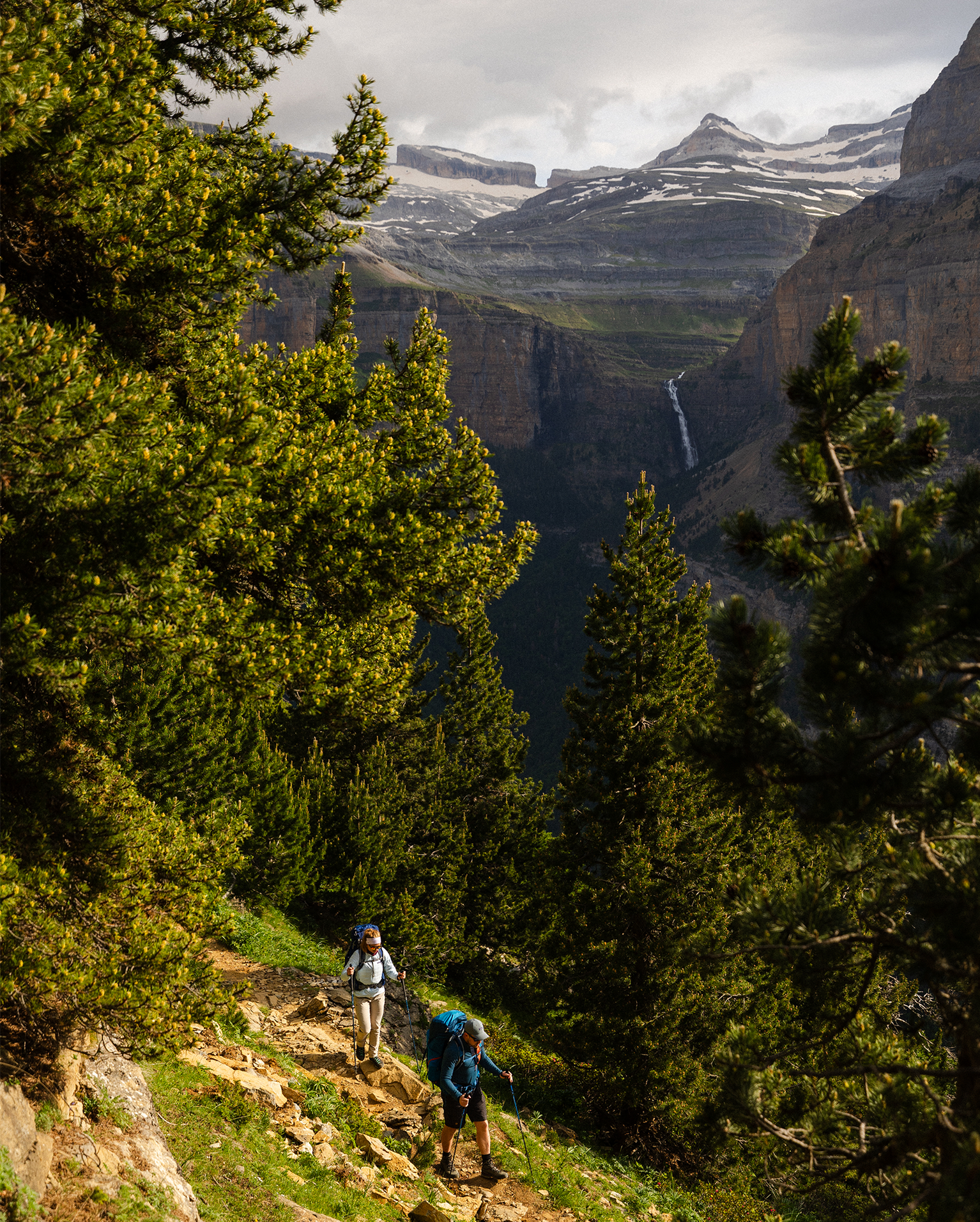 Two hikers on a lush mountain trail with pine trees, overlooking a valley and distant waterfall under a cloudy sky.