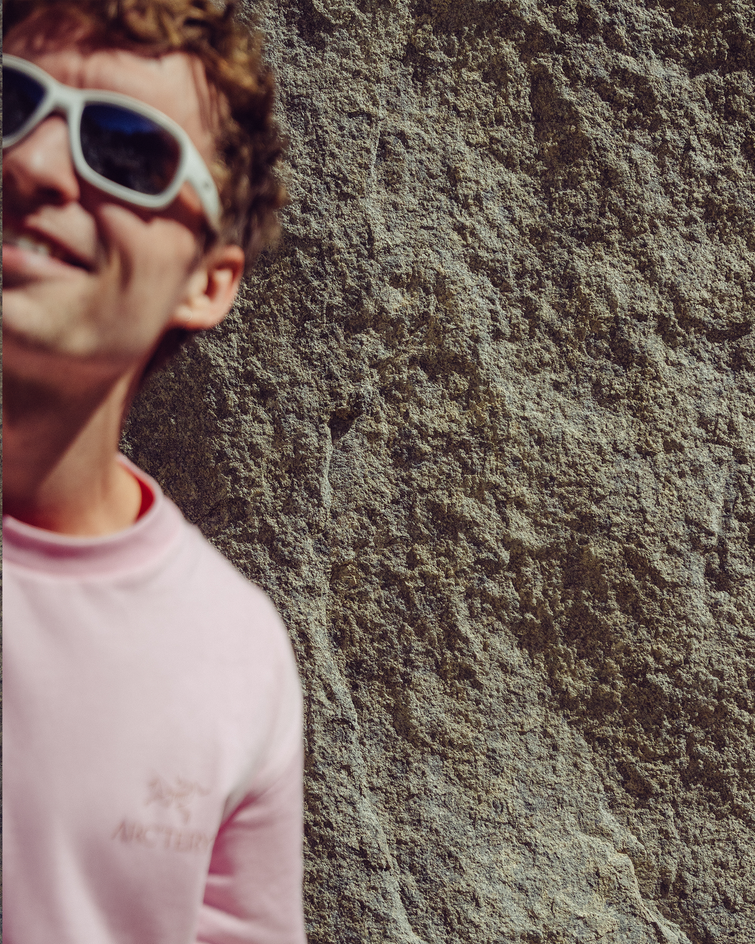 Person in pink shirt and white sunglasses smiling beside a textured rock wall.