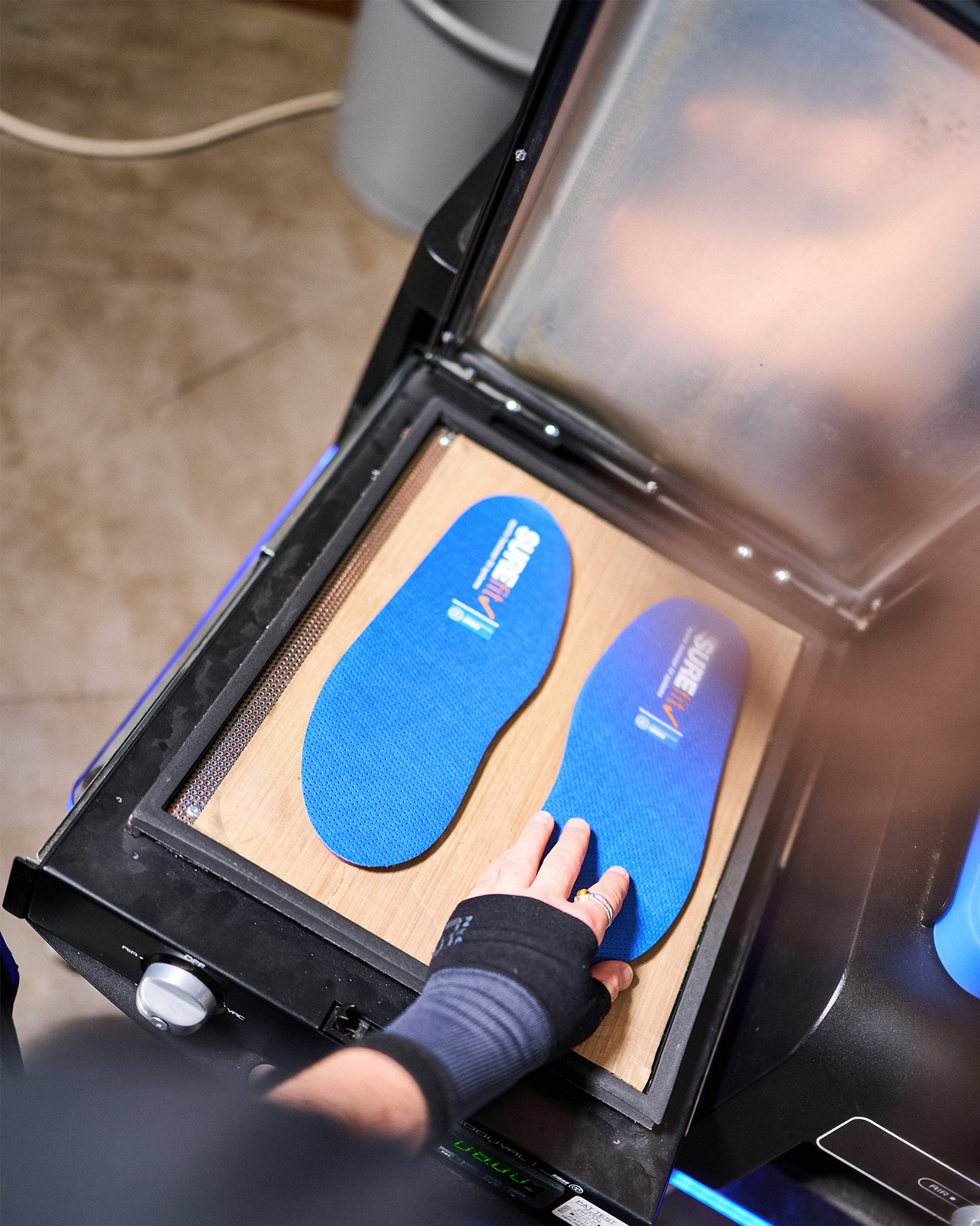 A person wearing a wrist brace places blue insoles into a machine for customization or heating.
