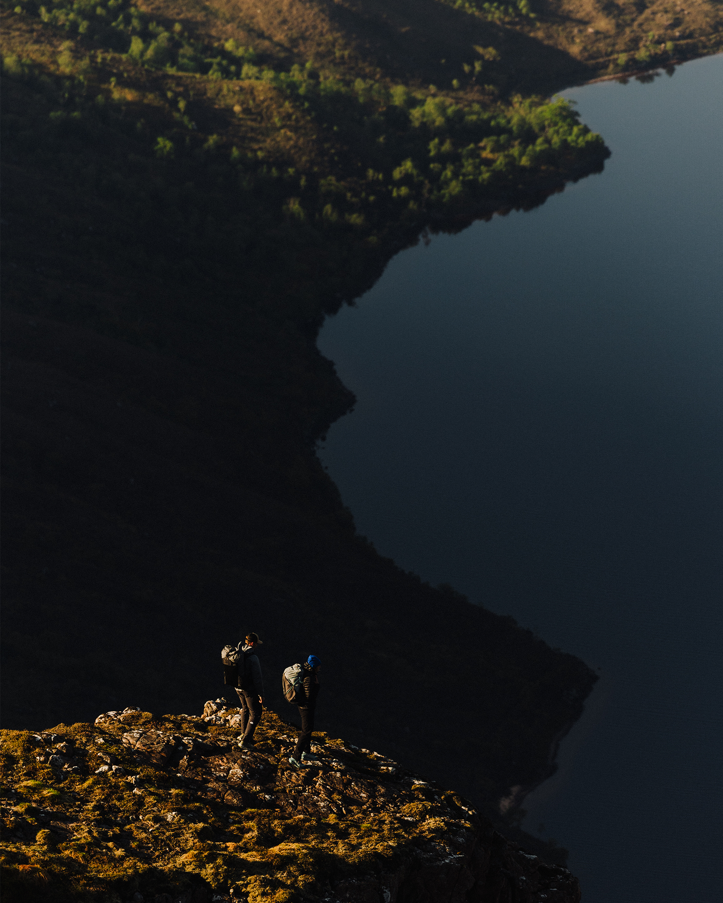 Two hikers stand on a rocky cliff, overlooking a serene lake and forest. The landscape is bathed in warm, golden light.