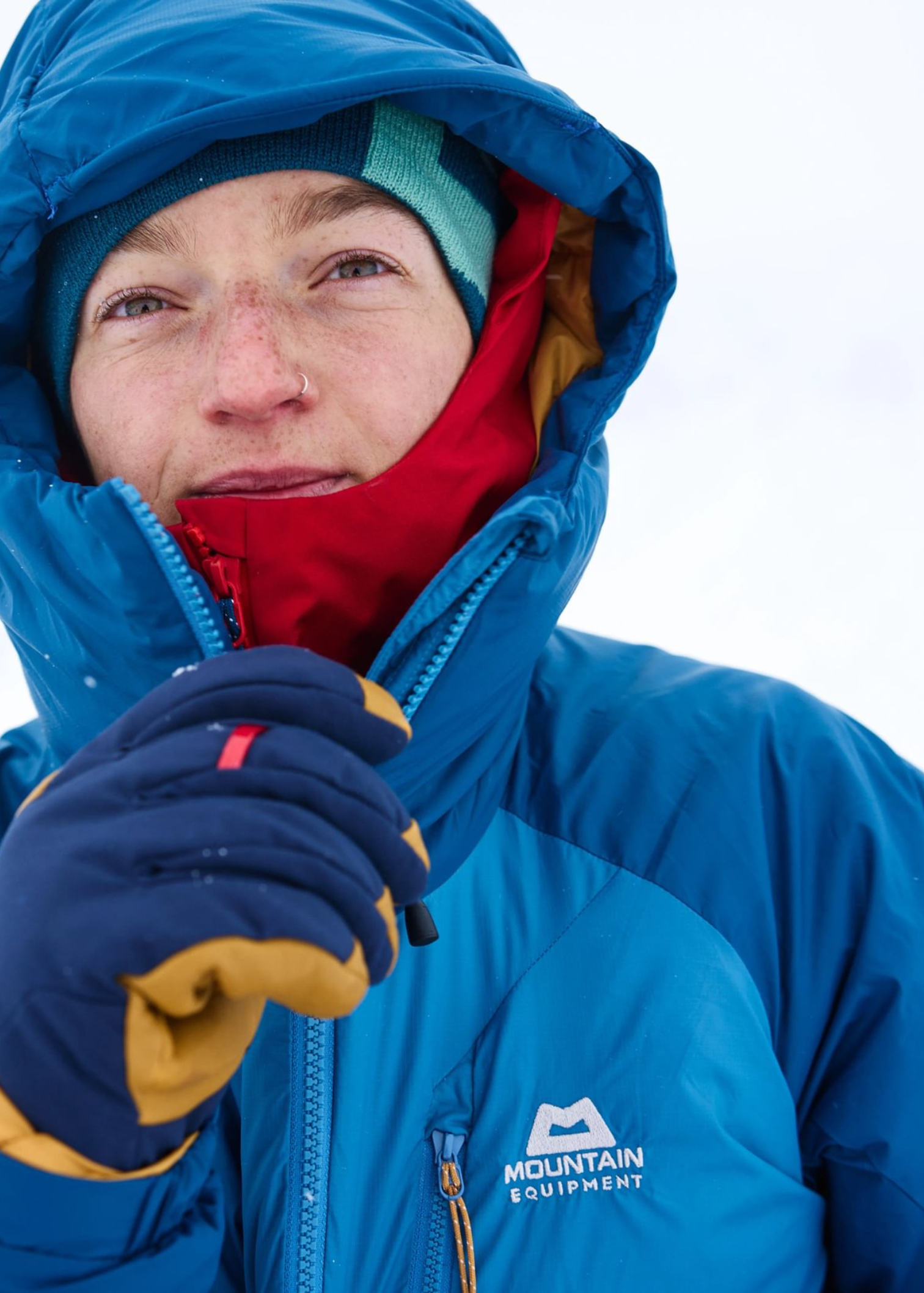 Person in a blue jacket and gloves, with a red inner layer, stands in a snowy setting, adjusting their hood.