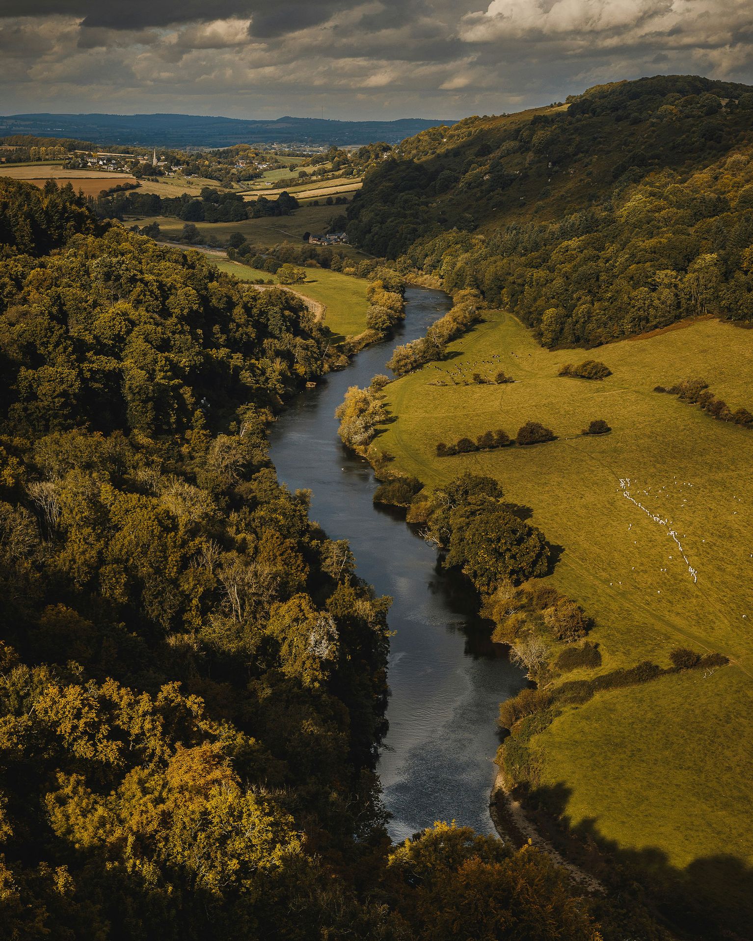 A winding river flows through lush, forested hills and green fields under a cloudy sky, capturing a serene landscape.