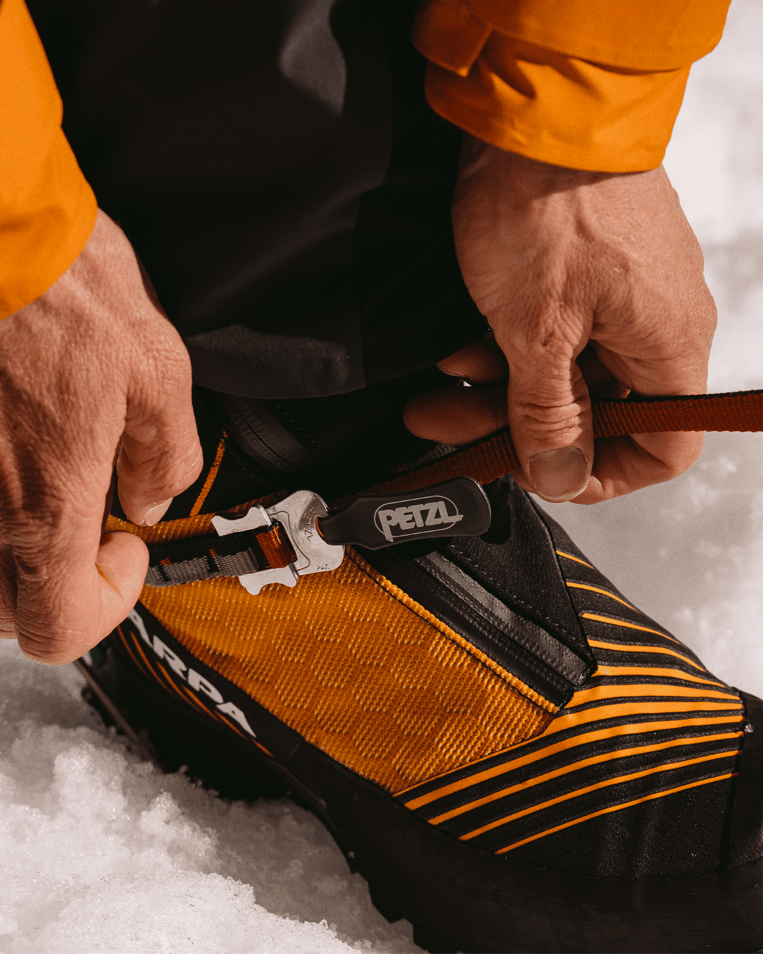 Close-up of hands adjusting a red strap on a yellow and black mountaineering boot in the snow.