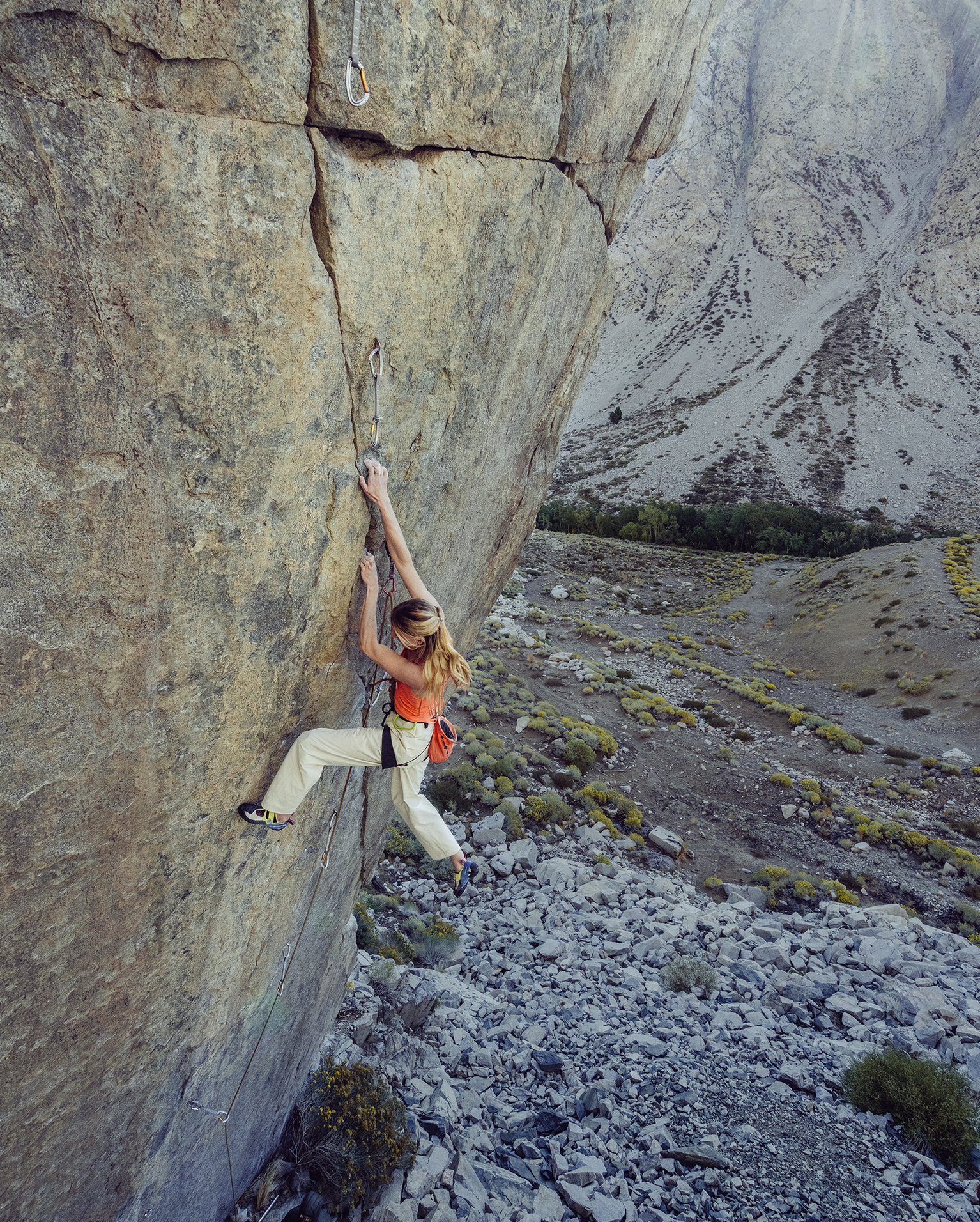 A woman climbs a steep rock face in a mountainous area, showcasing her skill and determination. She wears climbing gear and a red backpack.