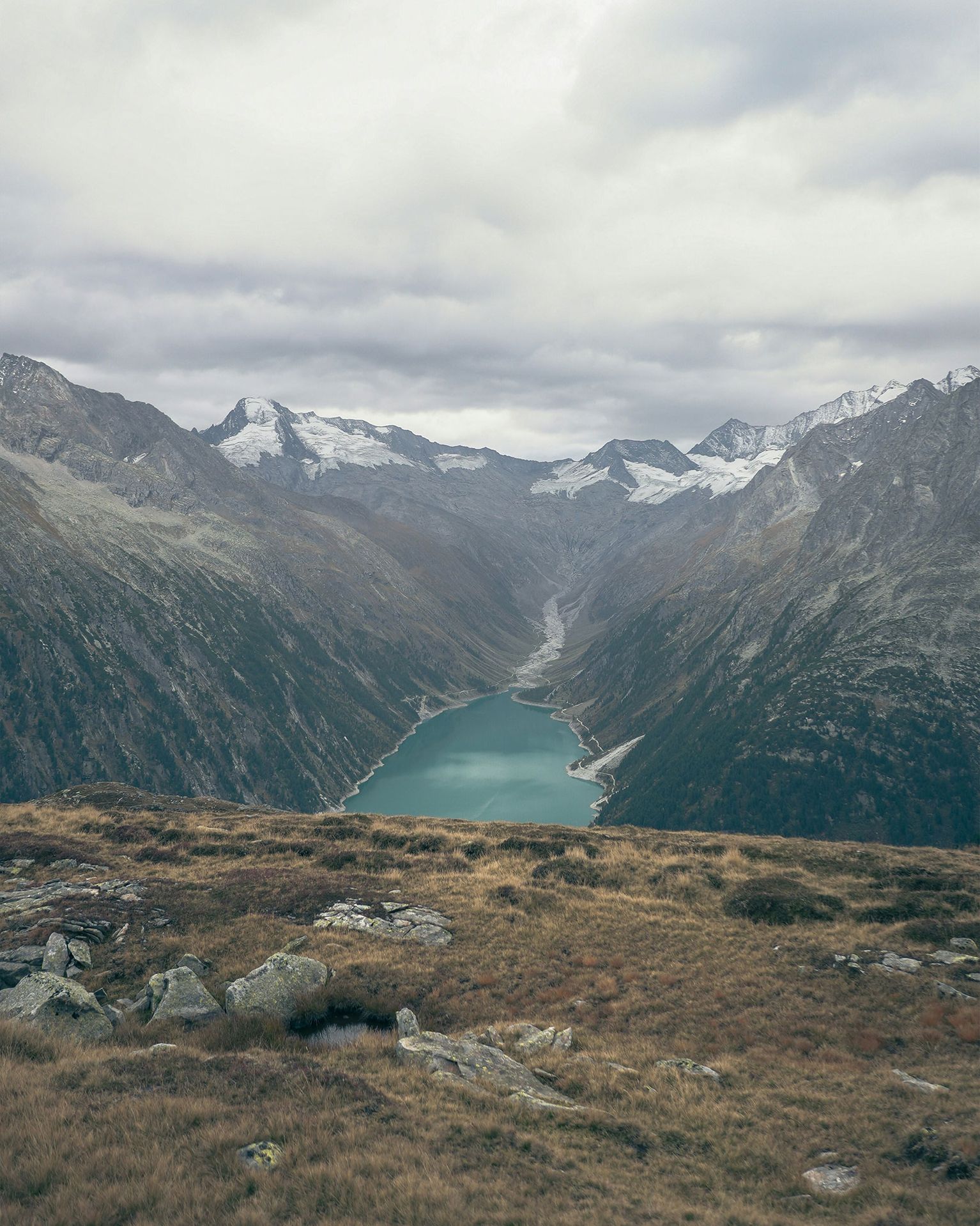 Mountain valley view with a turquoise lake surrounded by rugged peaks and cloudy sky. Grassy foreground with scattered rocks.