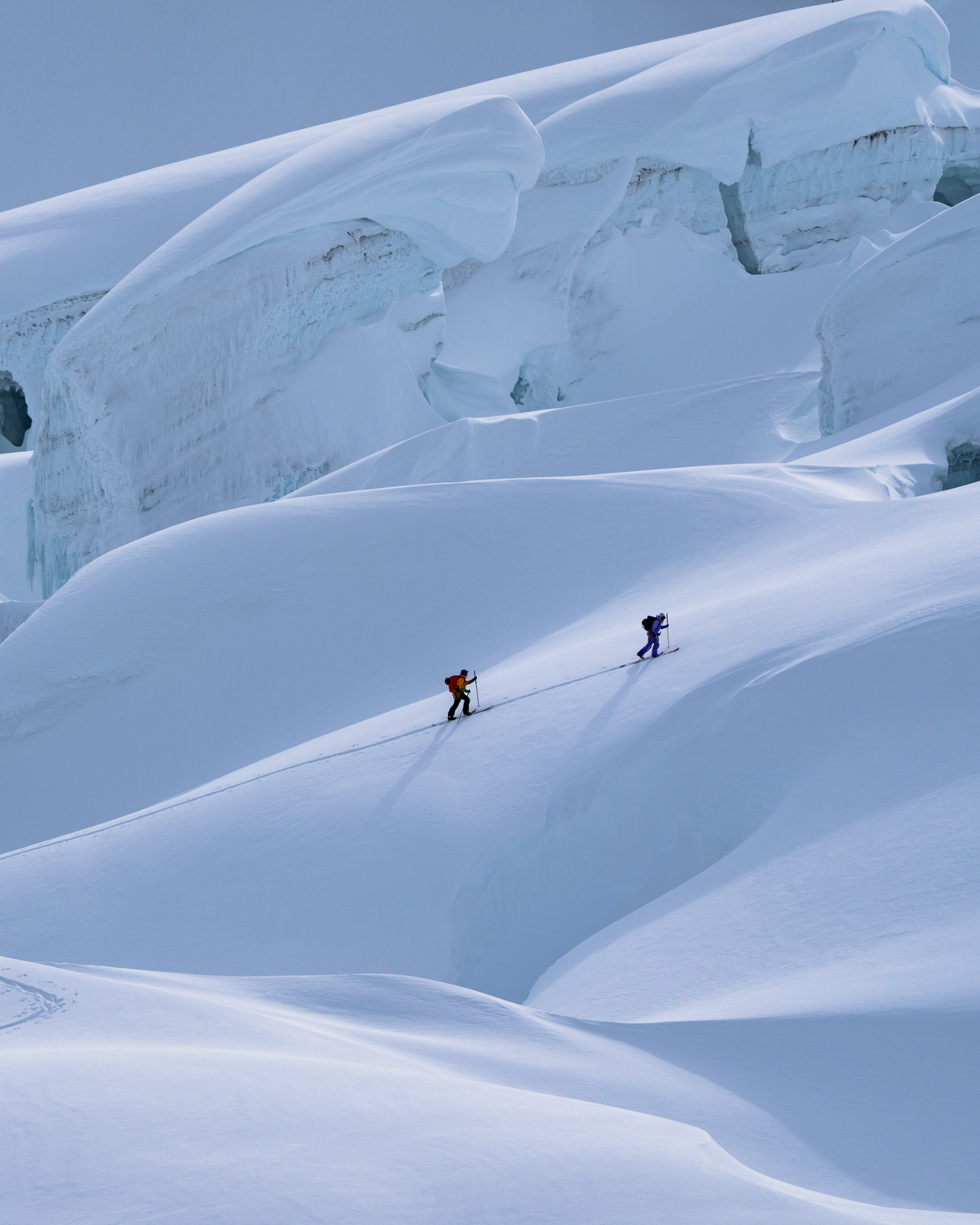 Two skiers traverse a snow-covered mountain slope with large ice formations in the background, under a clear blue sky.