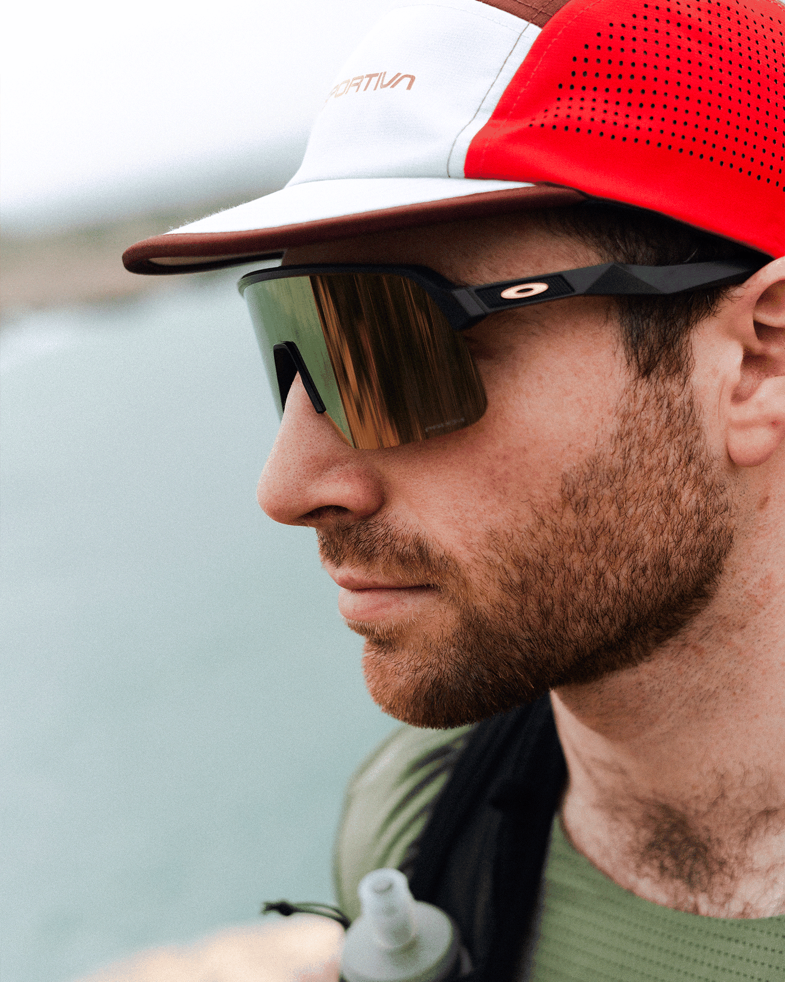 Man in a red and white cap wearing mirrored sunglasses and outdoor gear, standing near a body of water with a focused expression.