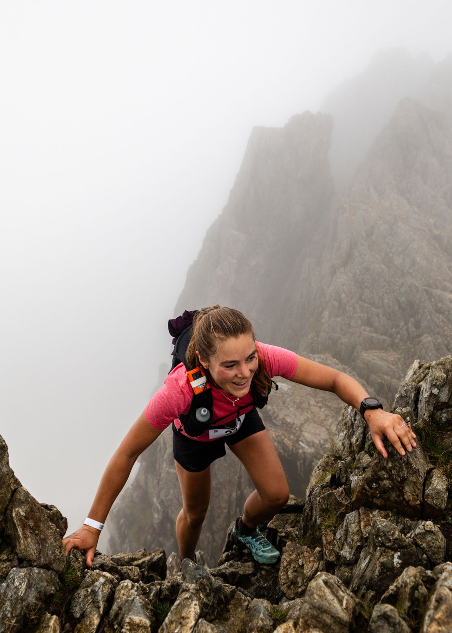 Person confidently climbing a rocky mountain trail, surrounded by mist. Wearing outdoor gear, including a red shirt and black shorts.