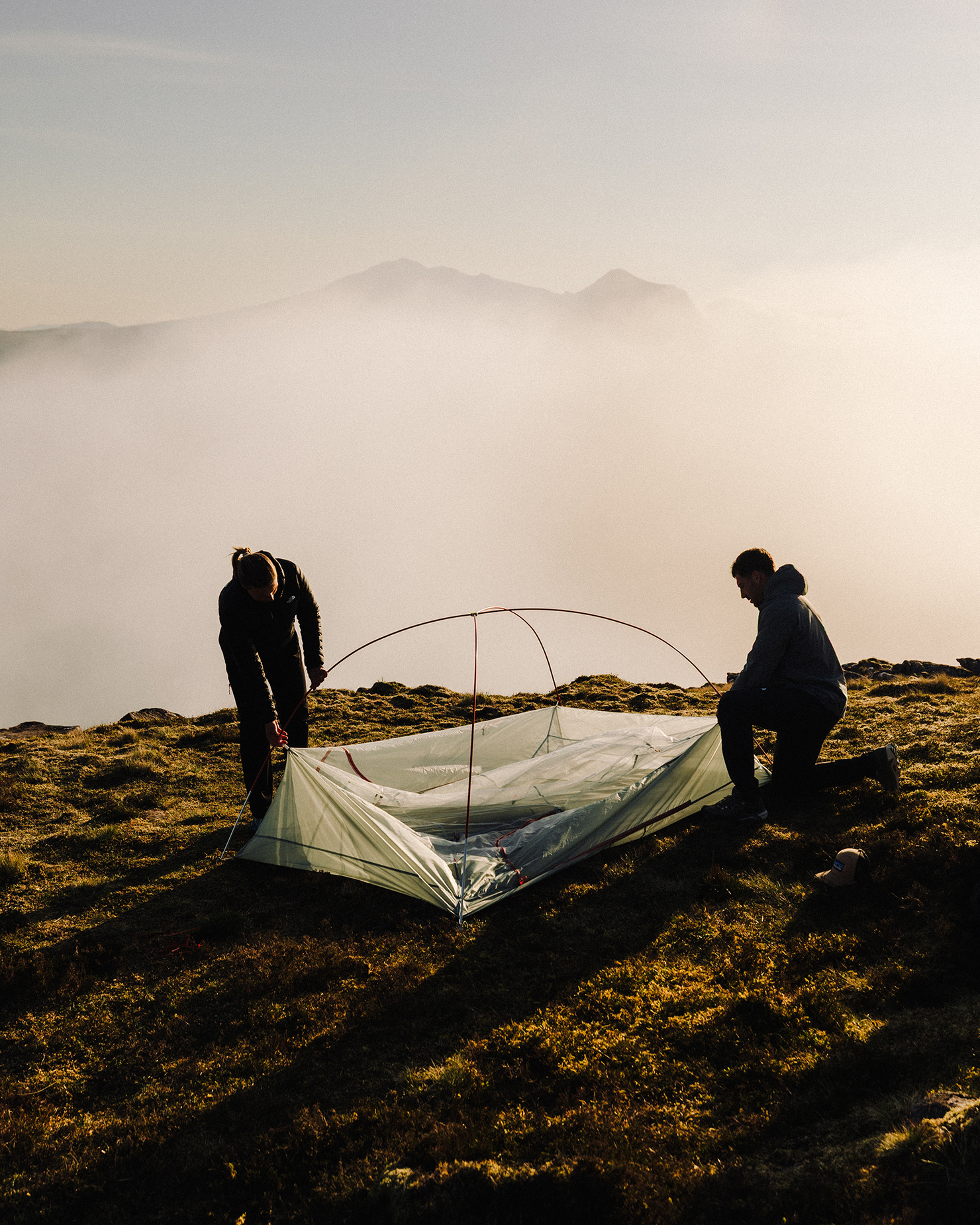 Two people setting up a tent on a mountaintop, surrounded by misty views and early morning light.