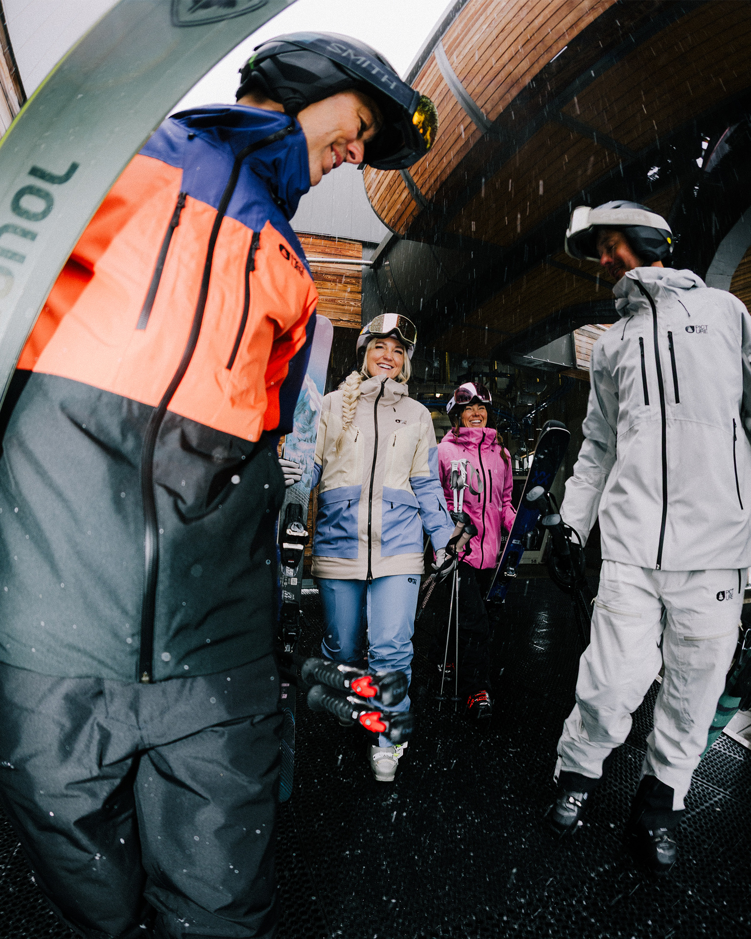 Group of skiers in colorful gear smiling and chatting under a wooden shelter, holding ski poles and wearing helmets.