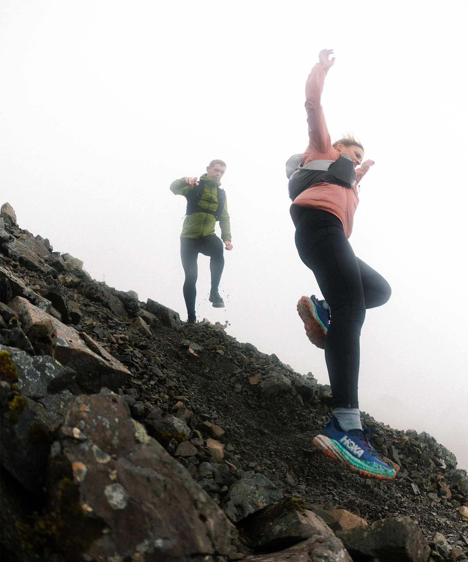 Two people trail running down a rocky, misty mountain slope, showcasing dynamic movement and vibrant athletic gear.