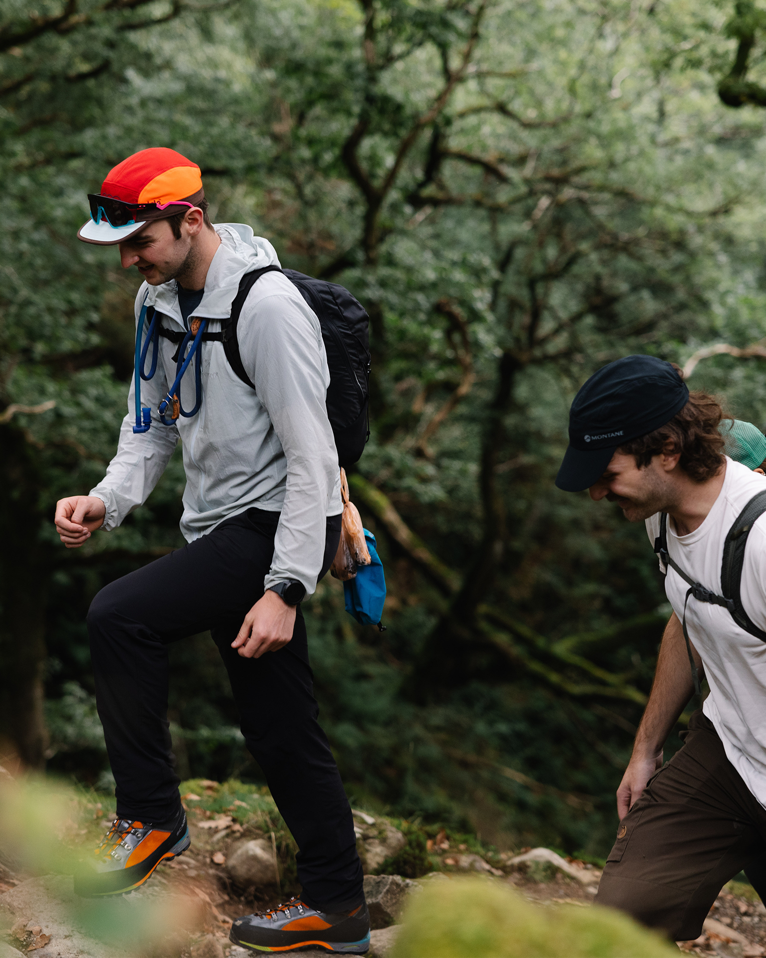 Two hikers with backpacks and caps traverse a forest trail, showcasing durable mountain sportswear and gear.