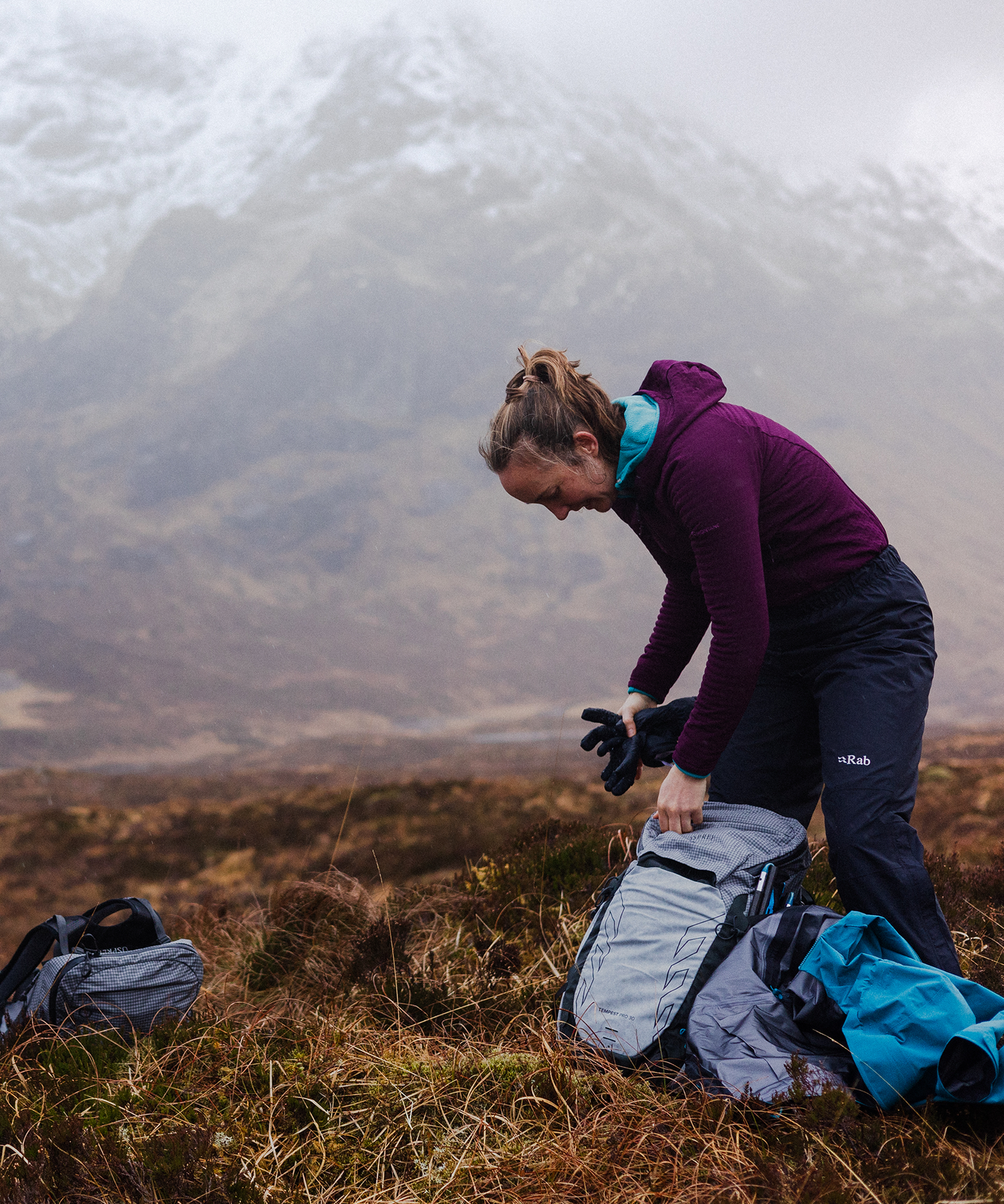 A person wearing mountain sports gear packs a backpack in a rugged, snowy mountain landscape.