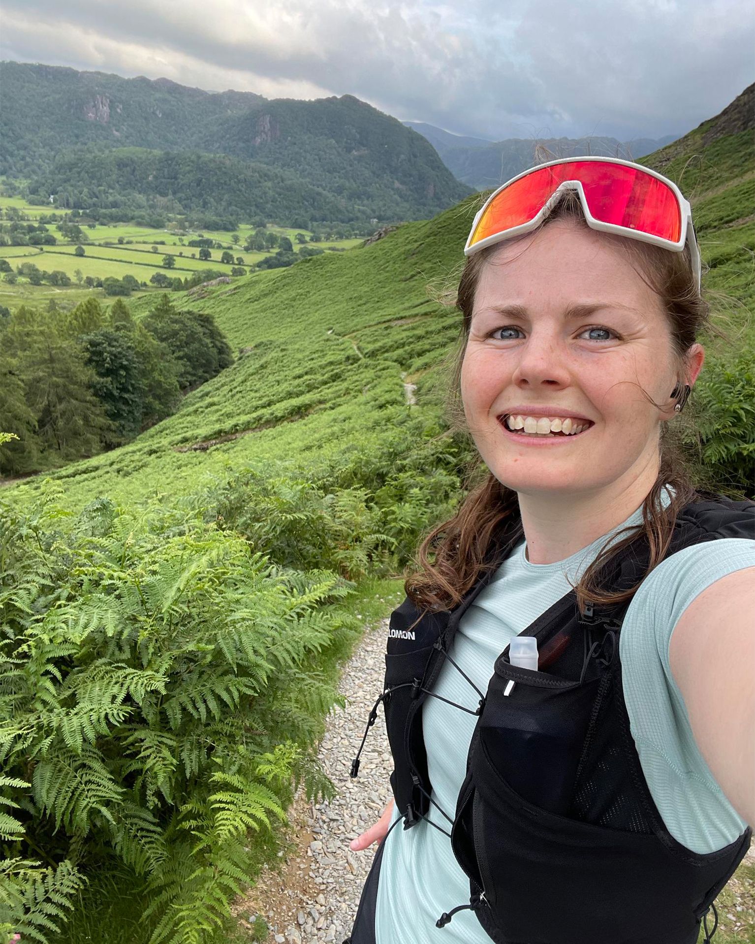 Person wearing sports gear smiling on a trail with lush greenery and hills in the background.