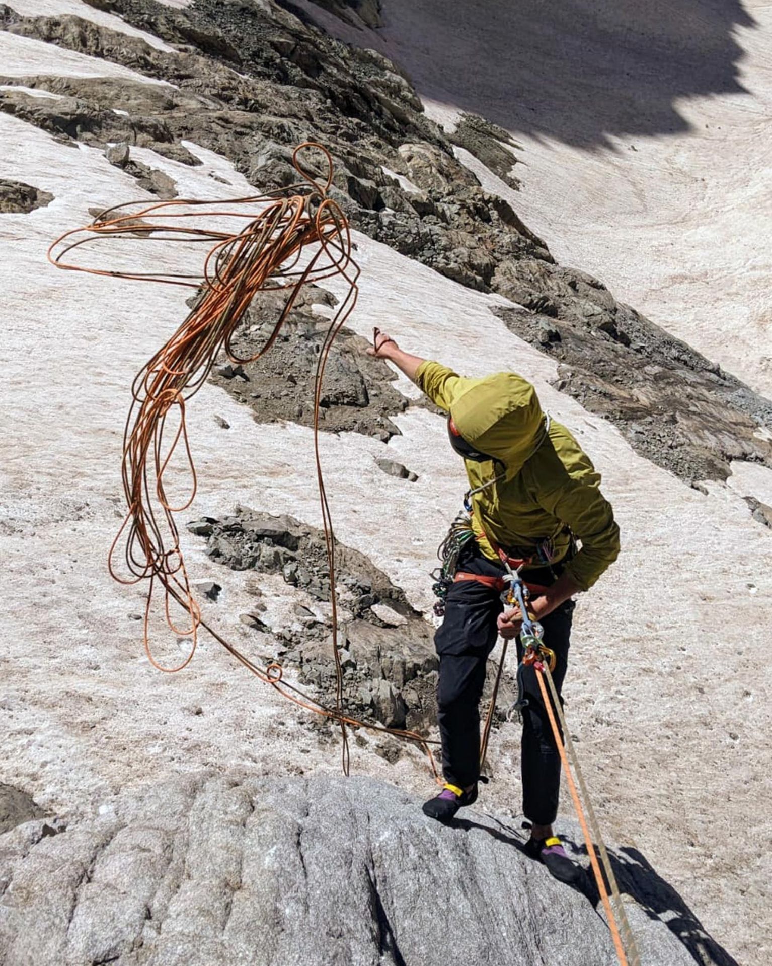 Climber in a green jacket throws a rope while standing on a rocky, snowy mountain slope.