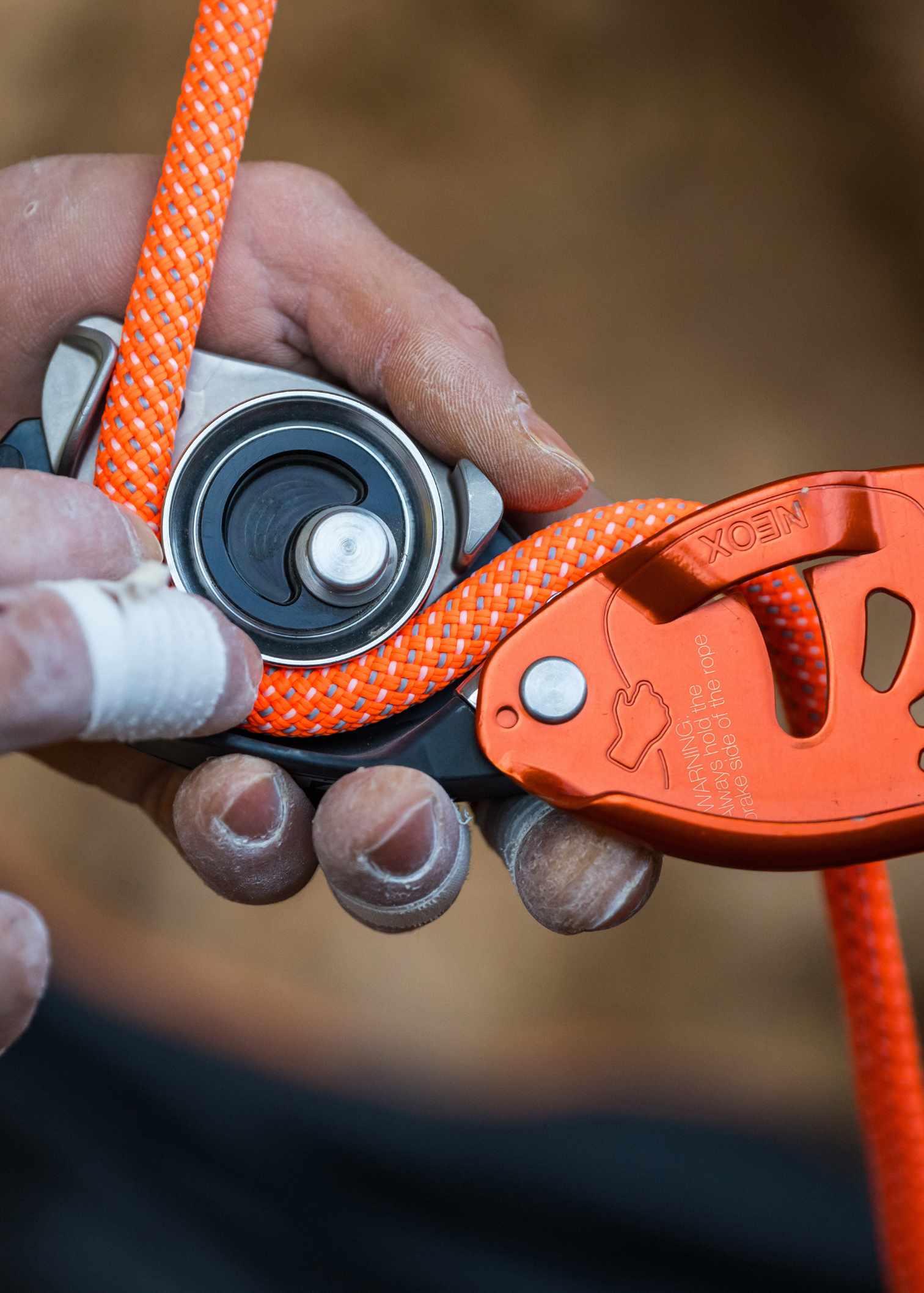 Close-up of hands using a belay device with orange climbing rope, fingers wrapped in climbing tape, demonstrating precise handling.