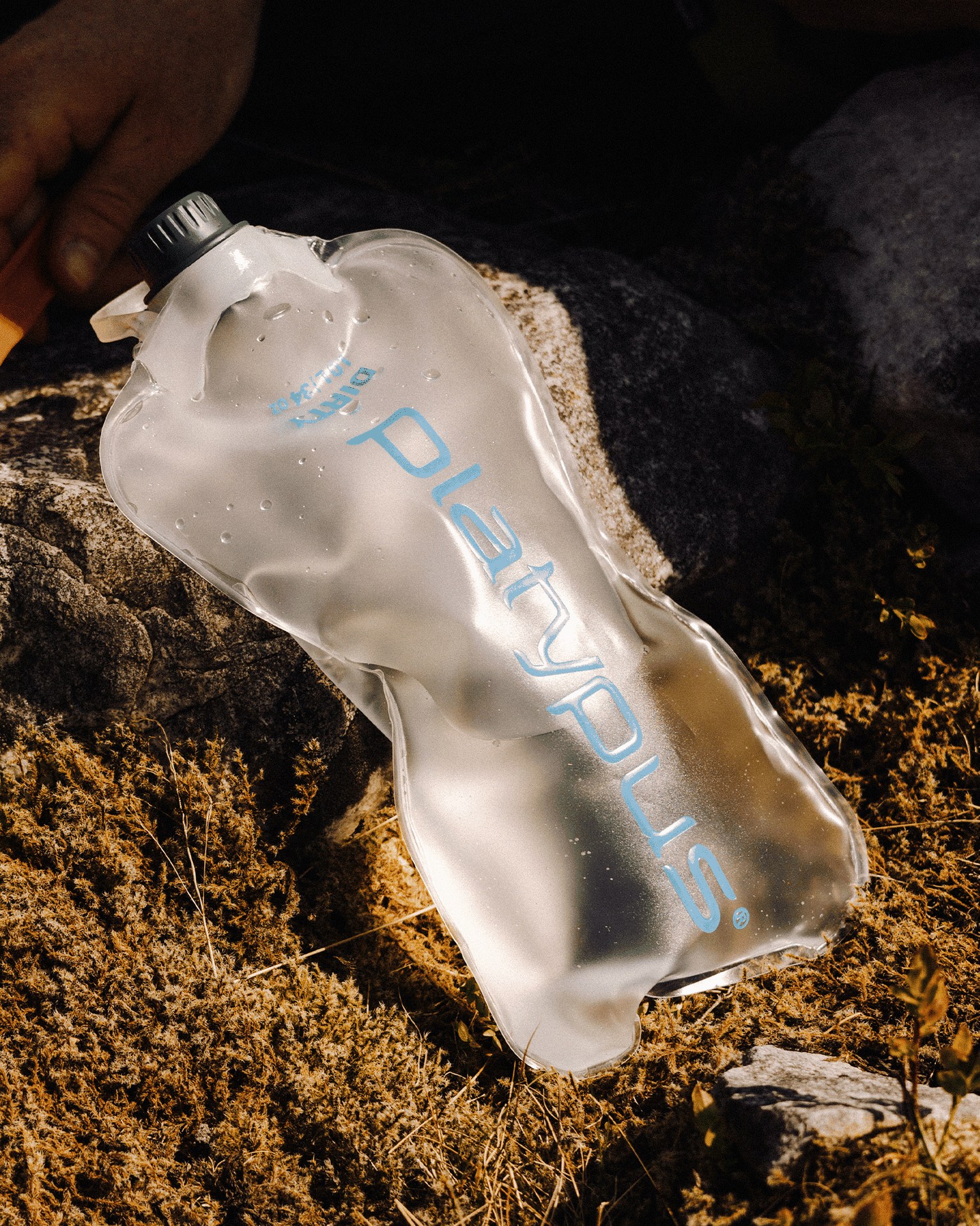 Flexible water bottle resting on the ground amidst rocks and grass, showing a durable design for outdoor use.