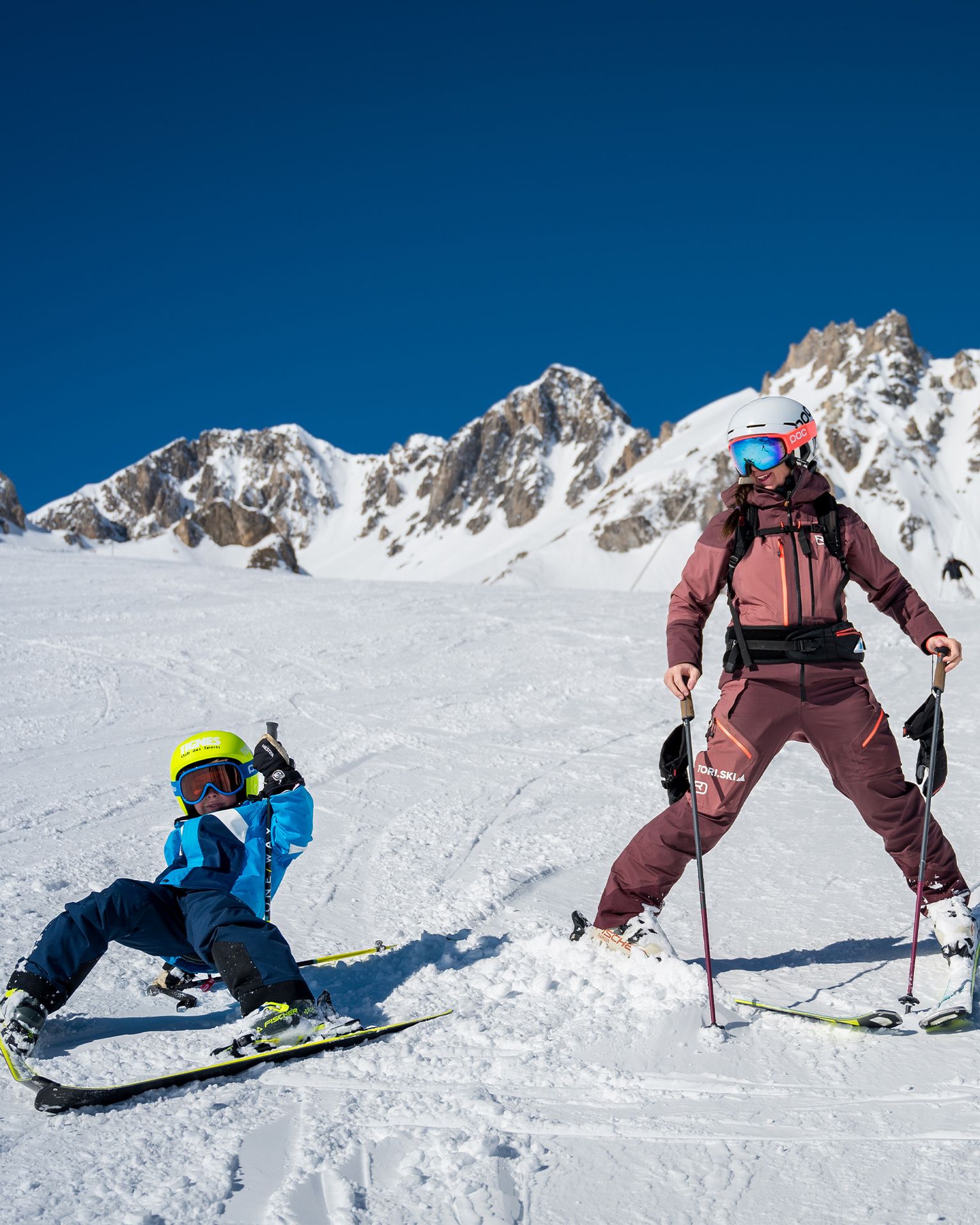 Adult and child skiing on a snowy mountain slope, surrounded by rugged peaks under a clear blue sky.