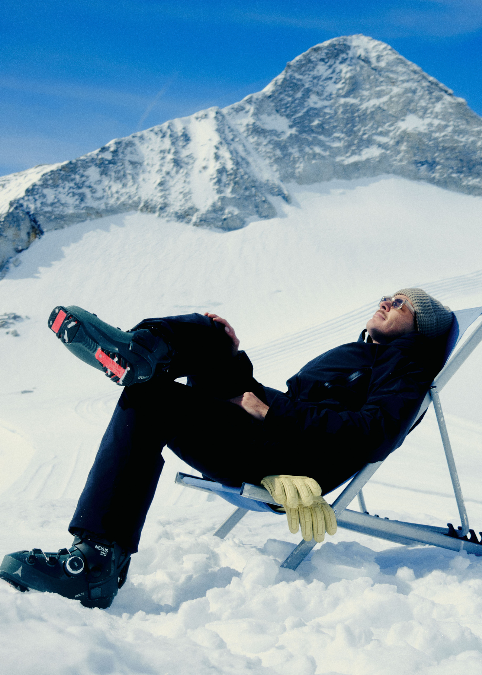 Skier reclines on a deck chair in the snow on a sunny day.