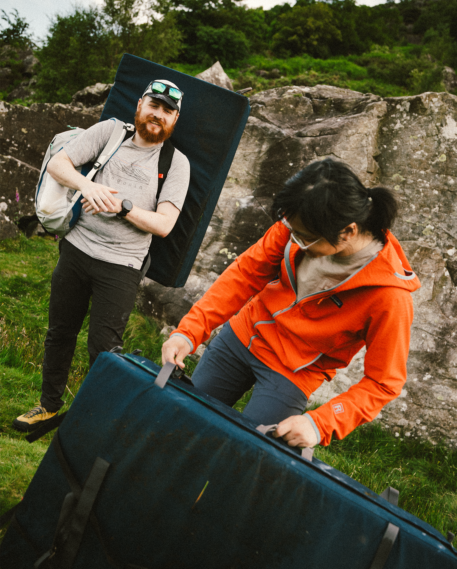 Two people prepare crash pads for bouldering in a rocky outdoor setting, displaying confidence and readiness for a climbing adventure.