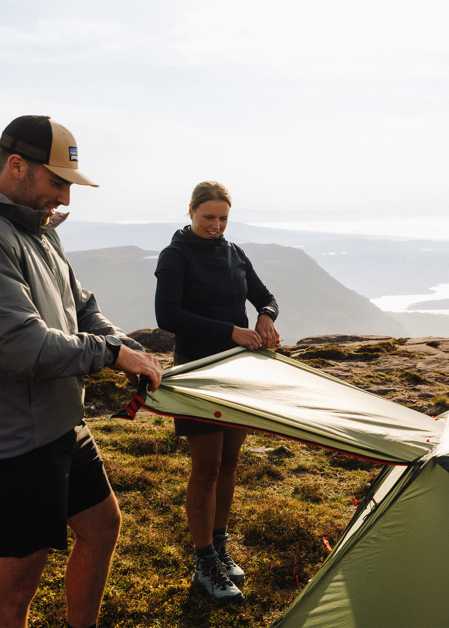 Two people setting up a green tent on a mountain with scenic views and distant water bodies.