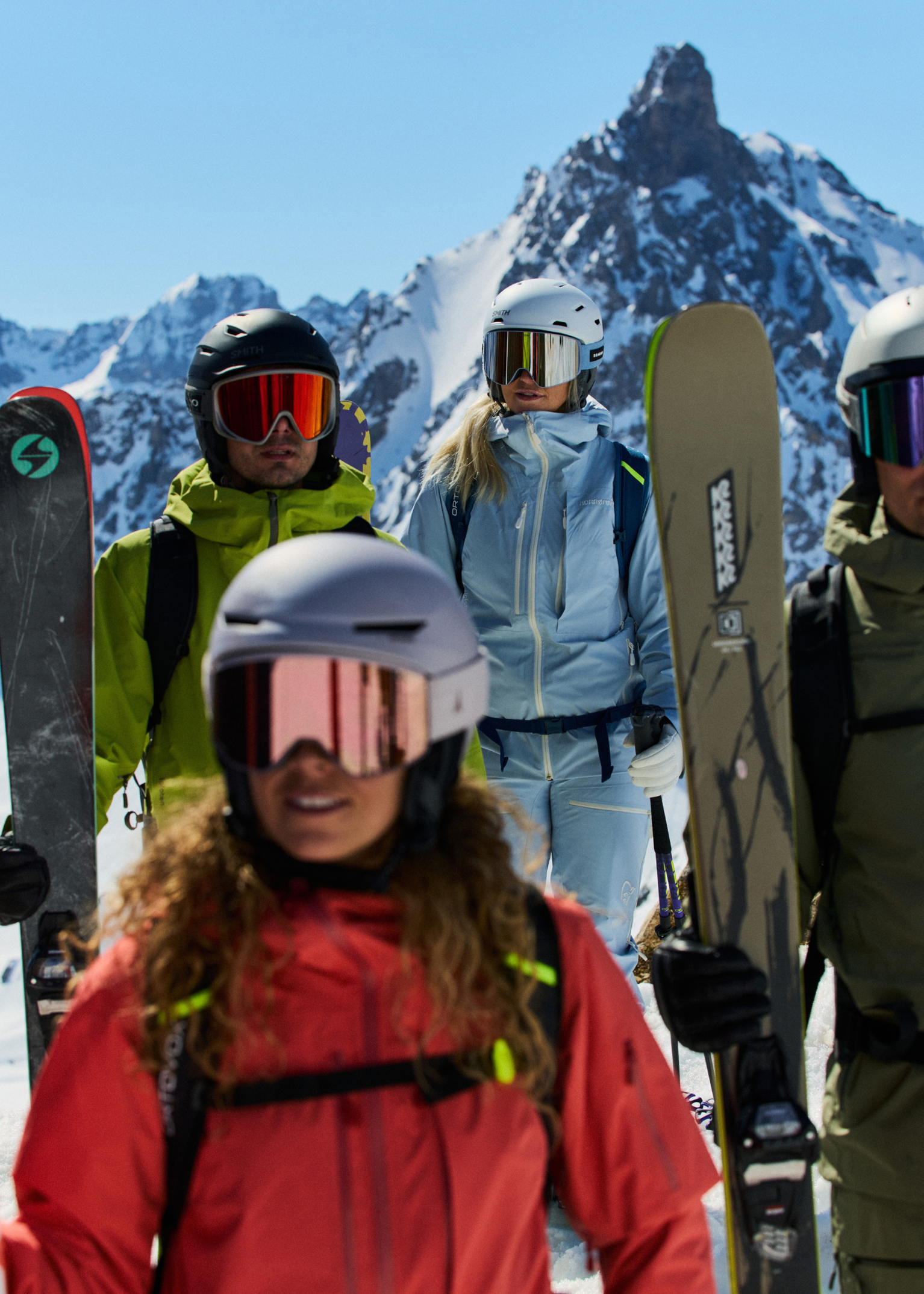 Skiers in vibrant gear stand on a snowy mountain, holding skis, with a majestic peak in the background under a clear blue sky.