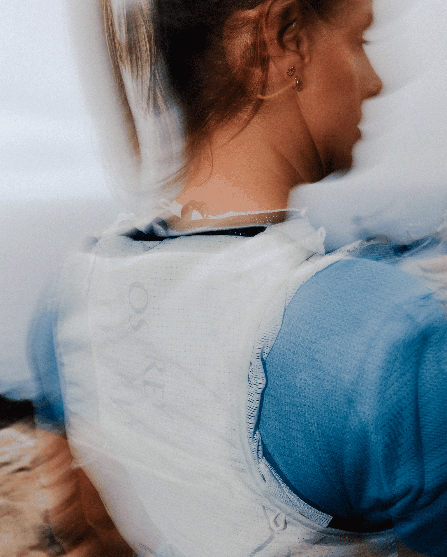 A person wearing a light blue top and white backpack, set against a blurred background, likely outdoors.