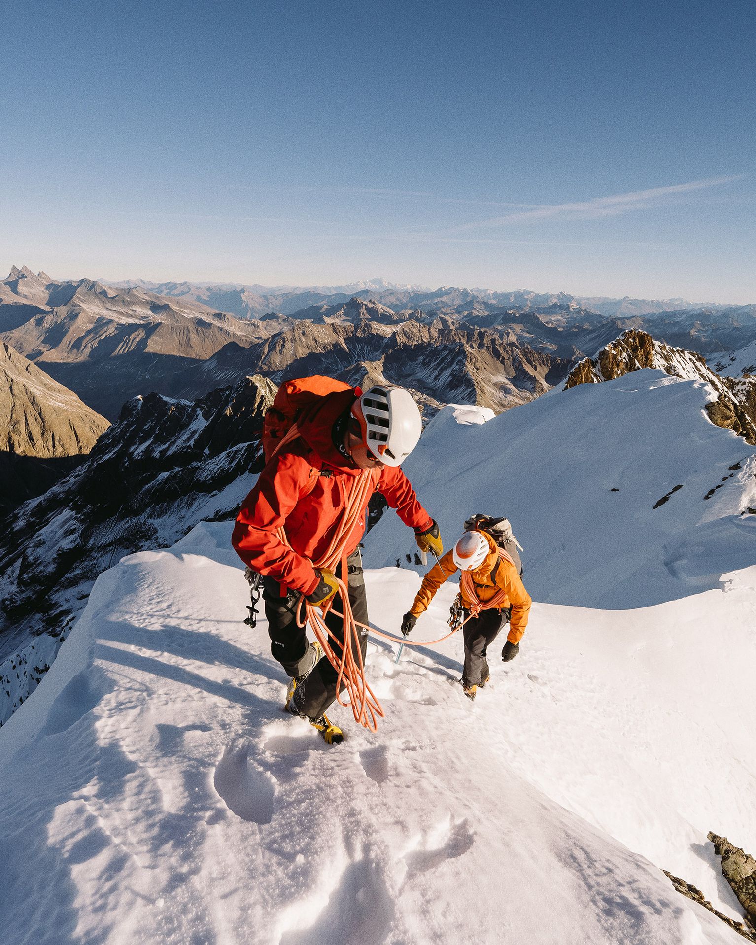Two climbers in helmets and gear ascend a snowy mountain peak under a clear blue sky, with vast mountain ranges in the background.